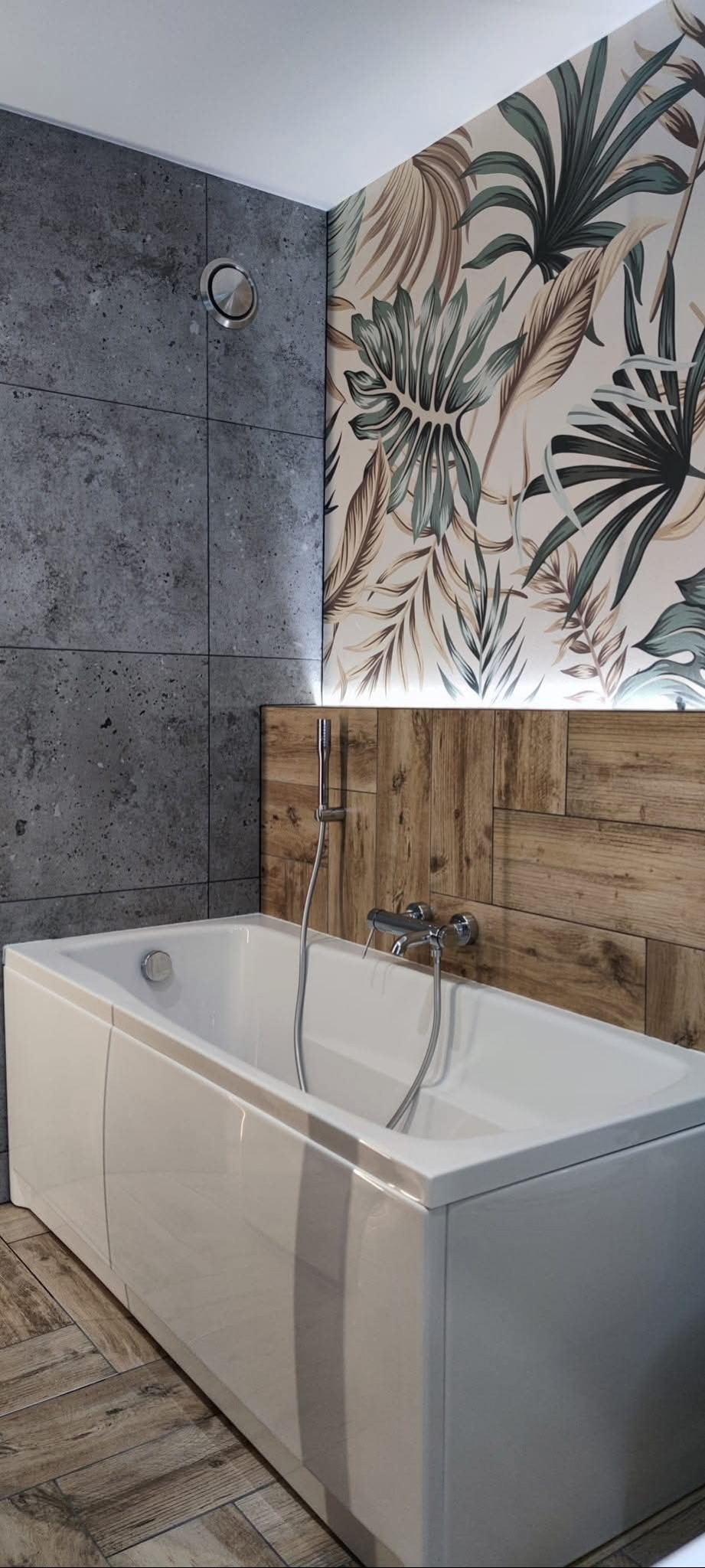 Nowoczesna łazienka z wanną, szarą ścianą z płytek i dekoracyjną tapetą w liście. Drewnopodobna okładzina i podłoga tworzą ciepły kontrast.