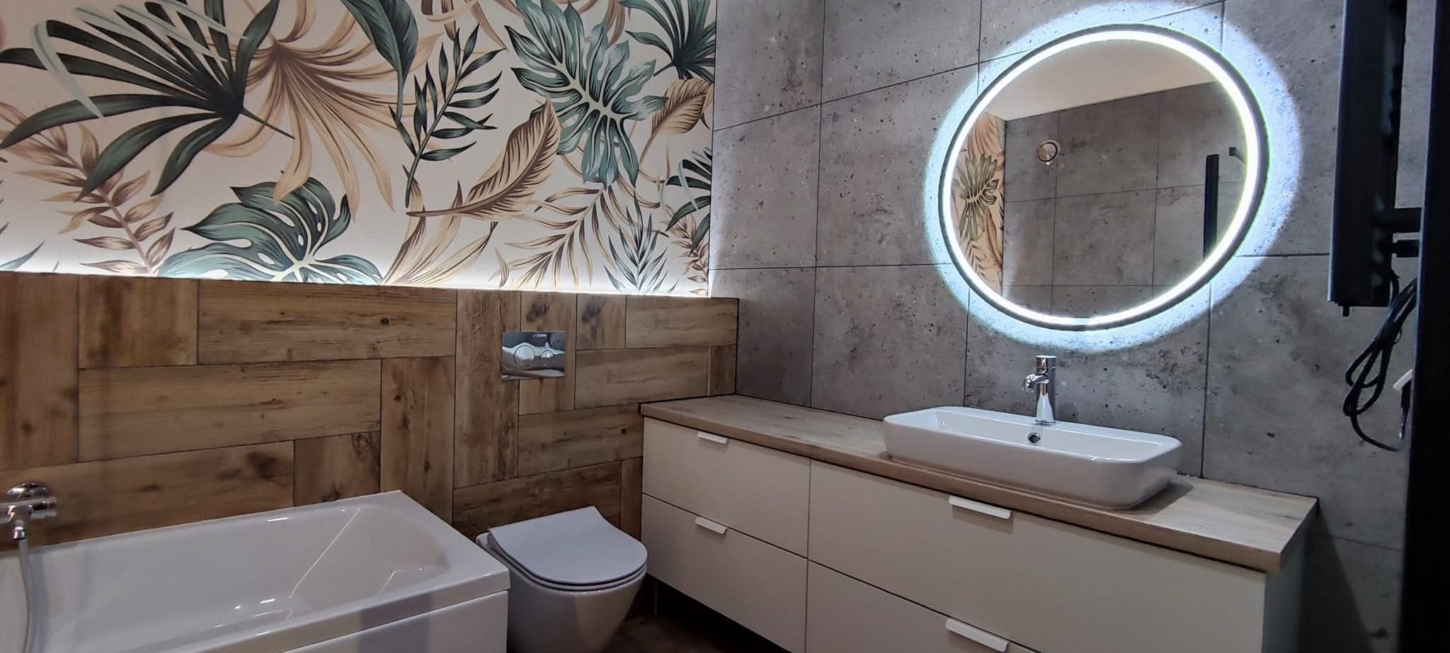 Nowoczesna łazienka z umywalką nablatową, okrągłym lustrem z podświetleniem LED, drewnianą boazerią i tapetą w tropikalne liście. Szare płytki imitujące beton.