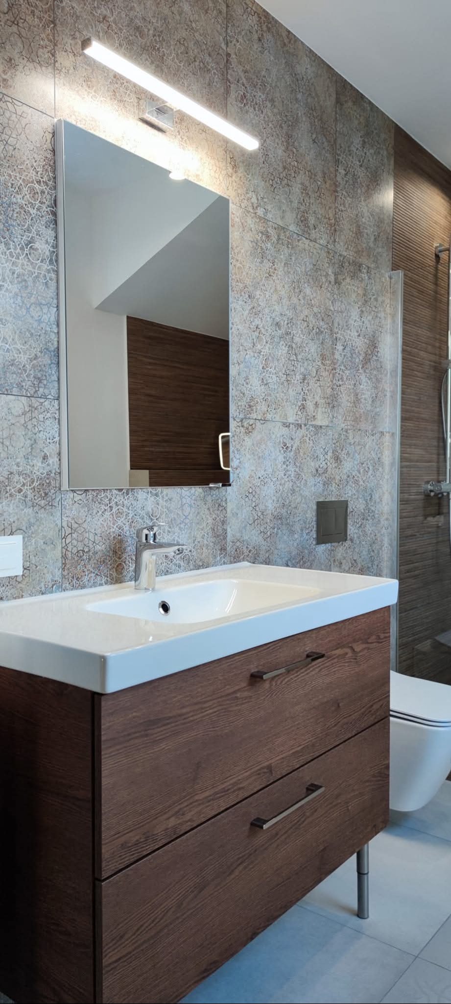 Nowoczesna łazienka z umywalką na szafce z ciemnego drewna, lustrem i oświetleniem LED. Płytki w odcieniach szarości i beżu z delikatnym wzorem. Kabina prysznicowa w tle.