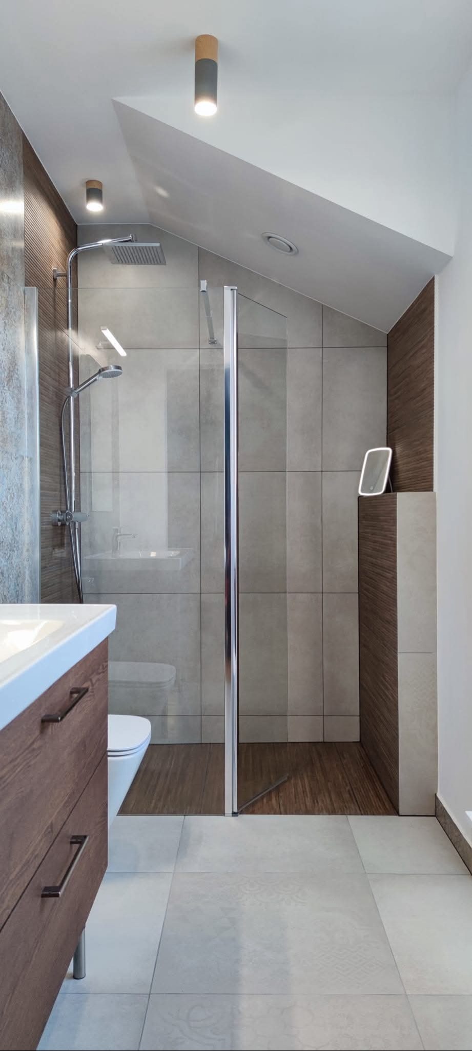 Nowoczesna łazienka na poddaszu z kabiną prysznicową typu walk-in, drewnianą szafką pod umywalką i oświetleniem punktowym. Uwagę zwraca skośny sufit.