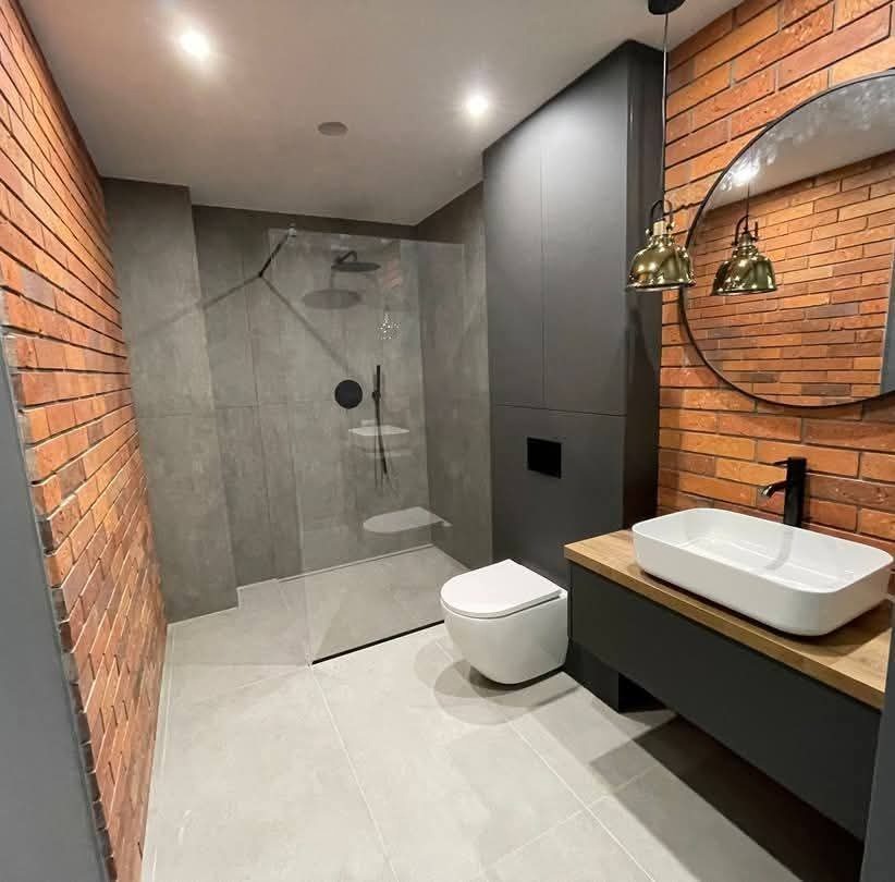 Nowoczesna łazienka z kabiną prysznicową typu walk-in, ceglaną ścianą i szarą zabudową. Umywalka nablatowa i okrągłe lustro dopełniają loftowy charakter.