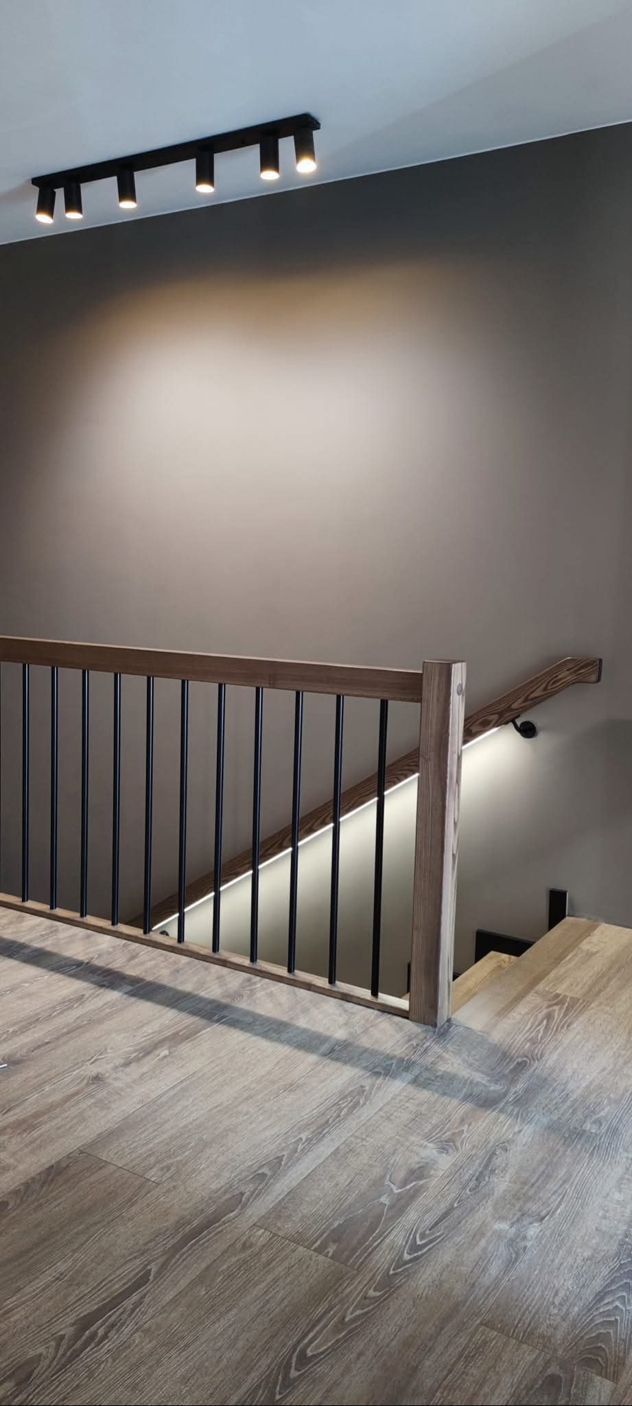 Nowoczesne schody z drewnianą balustradą i czarnymi szczeblami, oświetlone listwą LED. Na suficie reflektory punktowe. Podłoga wyłożona panelami imitującymi drewno.