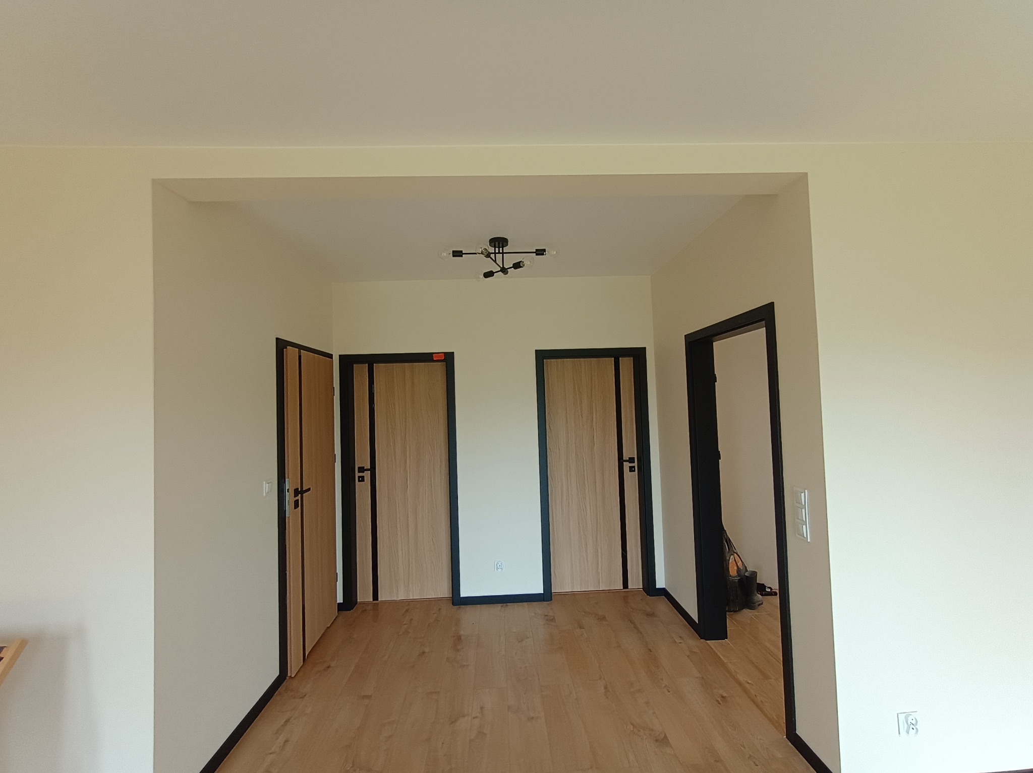 Jasne wnętrze po remoncie: panele podłogowe, pomalowane ściany w odcieniach beżu, dwoje drzwi z czarnymi ościeżnicami i nowoczesna lampa sufitowa.