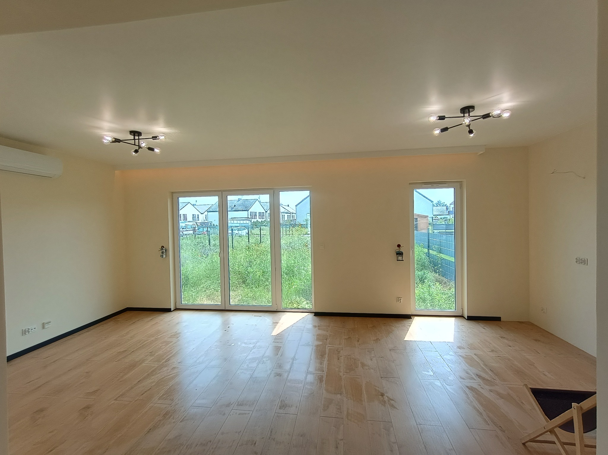 Jasne wnętrze z panelami podłogowymi imitującymi drewno, duże okna z widokiem na zieleń, nowoczesne lampy sufitowe. Minimalistyczny design, świeżo po remoncie.