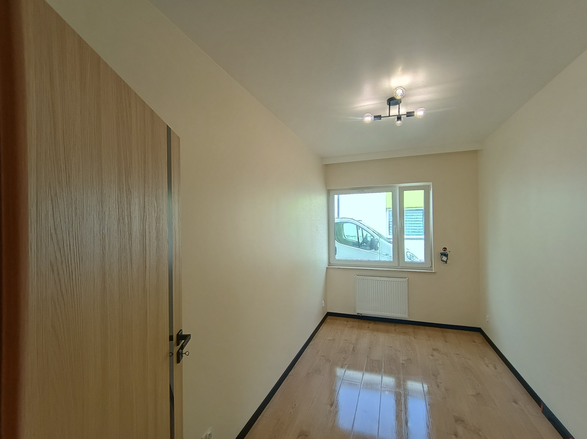 Jasne wnętrze po remoncie: parkiet, kremowe ściany, okno z widokiem na samochód. Nowoczesna lampa sufitowa i drewniane drzwi z czarną klamką.