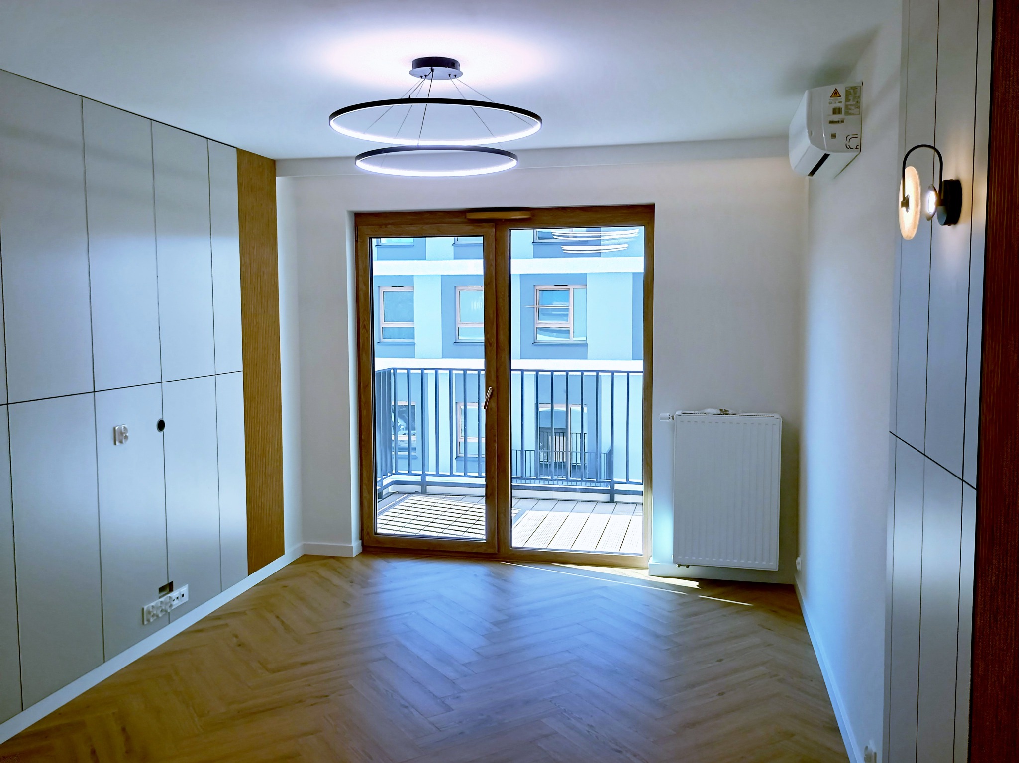 Jasne wnętrze po remoncie: parkiet w jodełkę, zabudowa szaf w kolorze złamanej bieli, okno balkonowe z widokiem na inne budynki, nowoczesne oświetlenie.