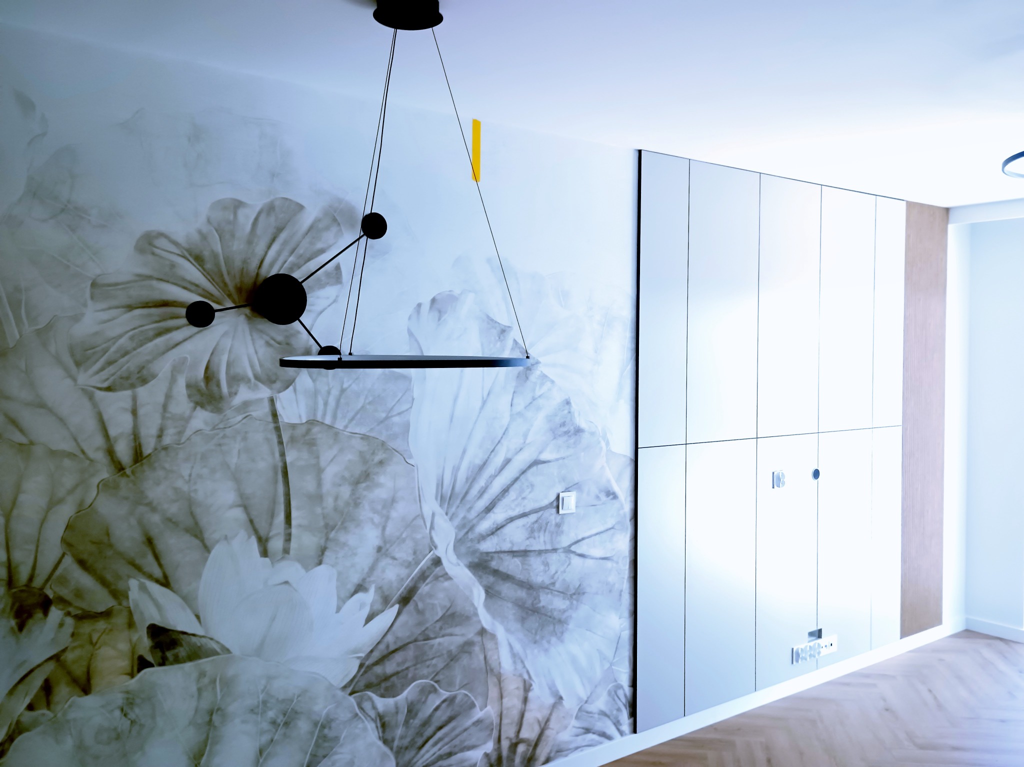 Nowoczesne wnętrze: ściana z motywem kwiatowym, minimalistyczna szafa w zabudowie i designerska lampa wisząca z żółtym akcentem. Parkiet ułożony w jodełkę.