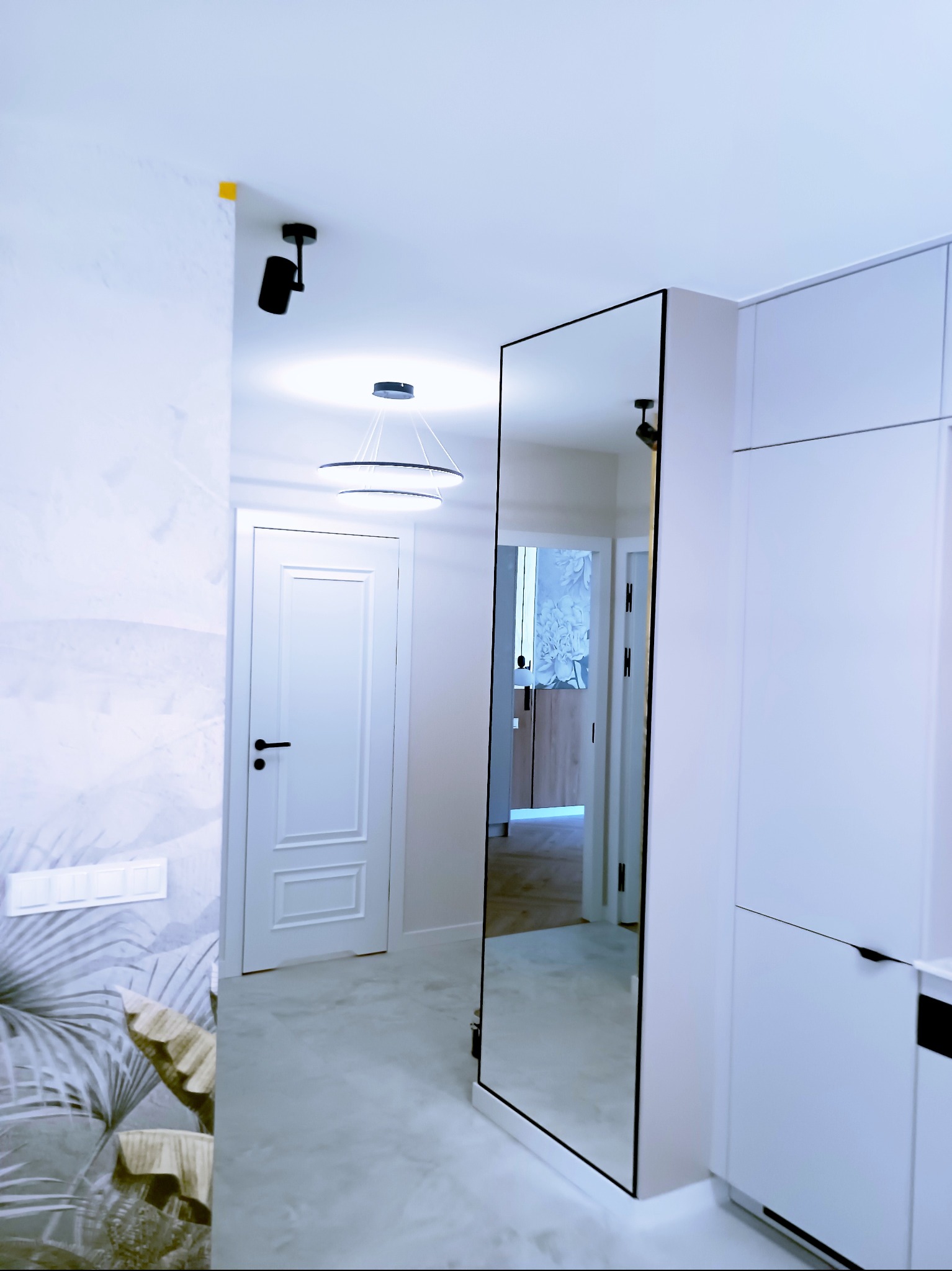 Nowoczesny przedpokój w bieli: lustro w czarnej ramie, drzwi z frezami, designerskie lampy sufitowe i tapeta w liście. Minimalistyczny i elegancki design.