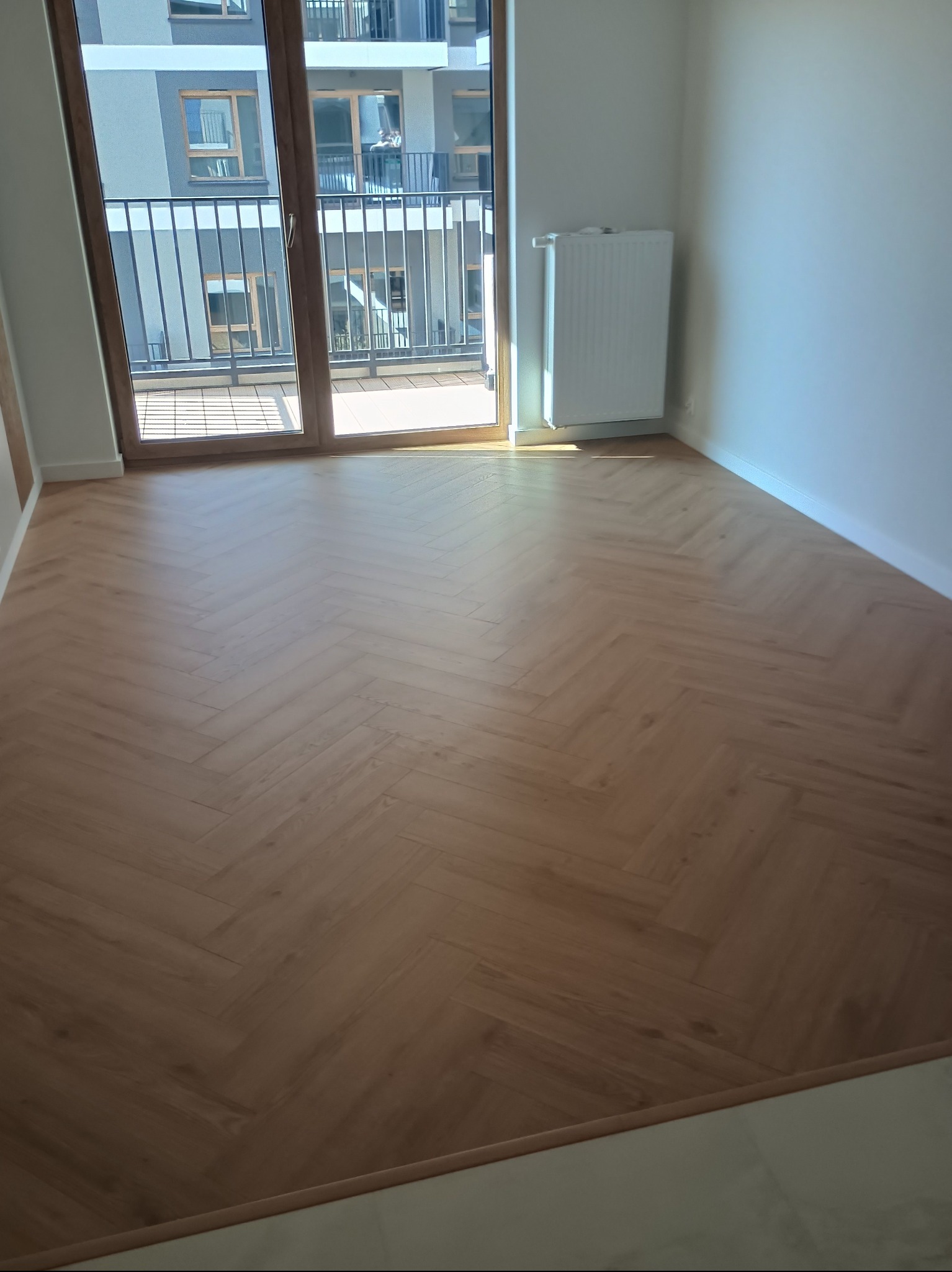 Ułożone panele podłogowe w jodełkę w jasnym, nowoczesnym pokoju z balkonem i grzejnikiem. Widok na budynek z zewnątrz przez otwarte drzwi balkonowe.
