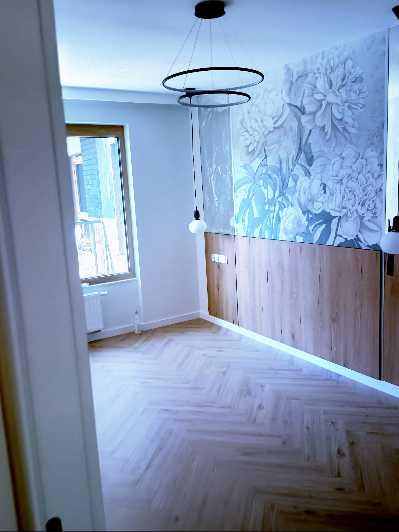 Jasne panele ułożone w jodełkę, szafka z podświetleniem LED i fototapeta z motywem kwiatowym w pokoju z oknem. Nowoczesne lampy wiszące.