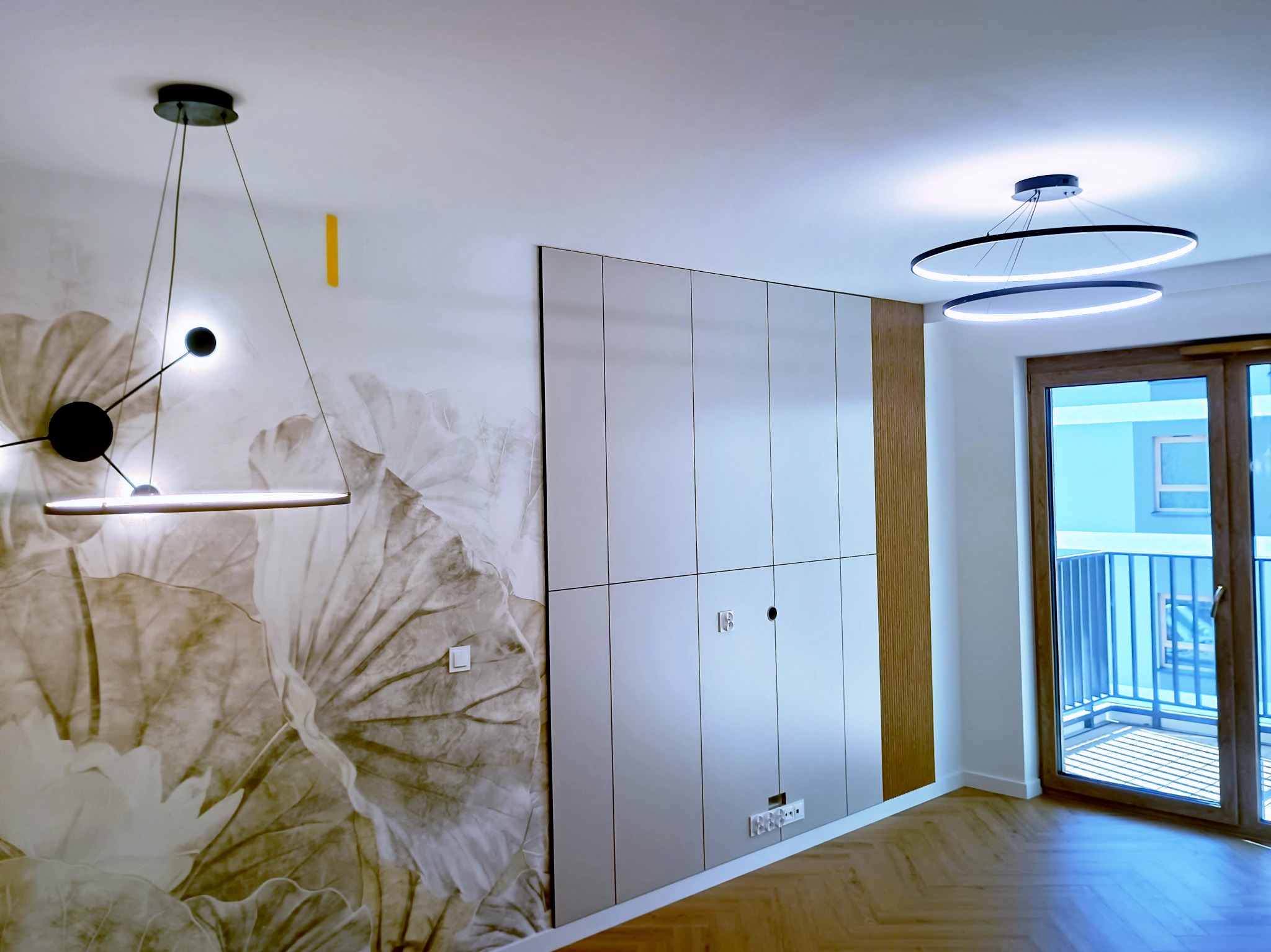 Nowoczesne wnętrze: ściana z fototapetą w kwiaty, zabudowa meblowa z gniazdkami, parkiet w jodełkę, okno balkonowe i designerskie lampy sufitowe.