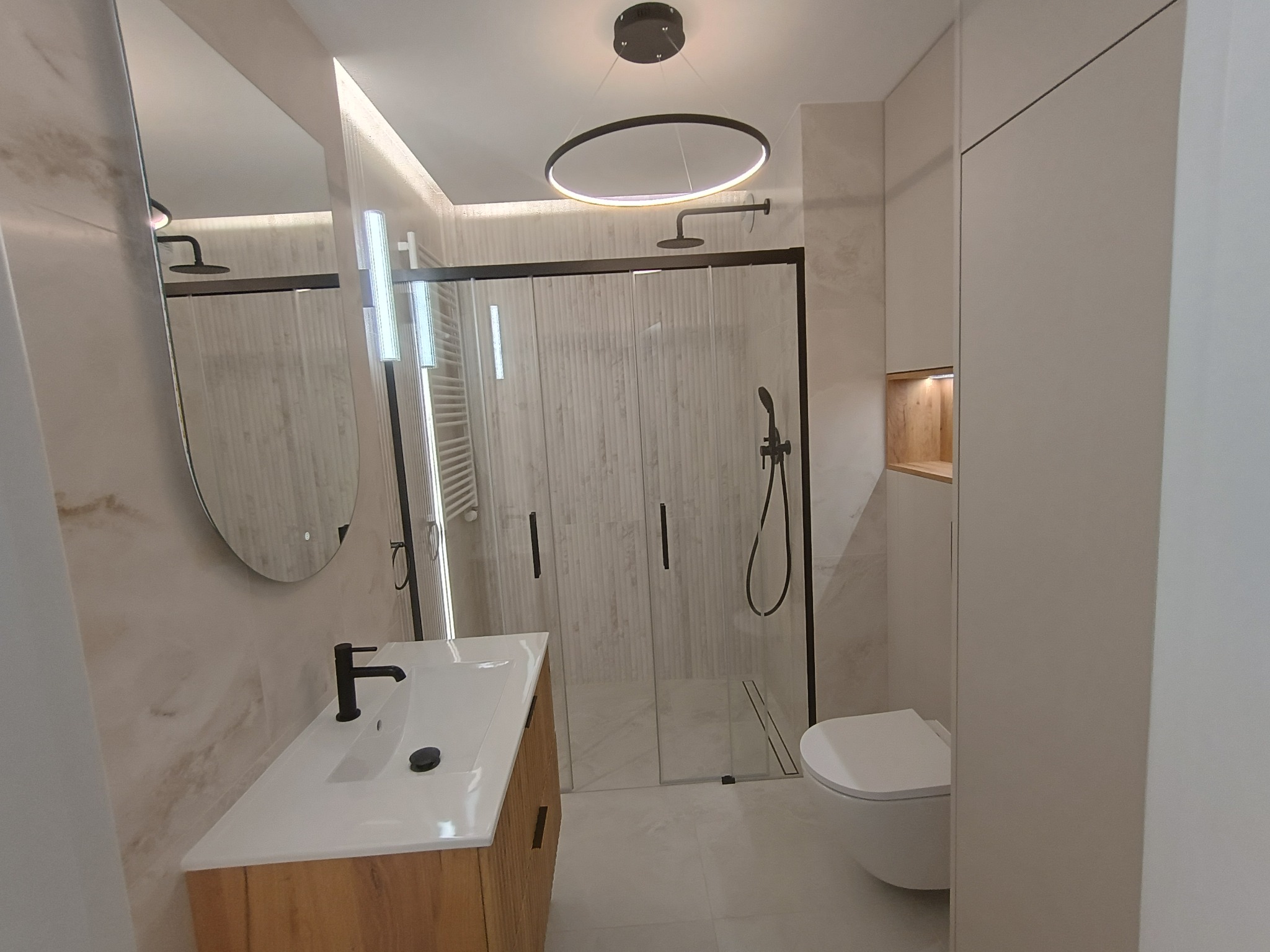 Nowoczesna łazienka z kabiną prysznicową typu walk-in, umywalką z drewnianą szafką i okrągłą lampą sufitową. Minimalistyczny design, jasne kolory i marmurowe wykończenie.