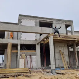 Surowy stan dwupiętrowego domu z betonu komórkowego w trakcie budowy, z osobą w masce stojącą na konstrukcji i rozkładającą ręce, widoczny zarys przyszłych okien i drzwi, drewniane podpory i szalunki.