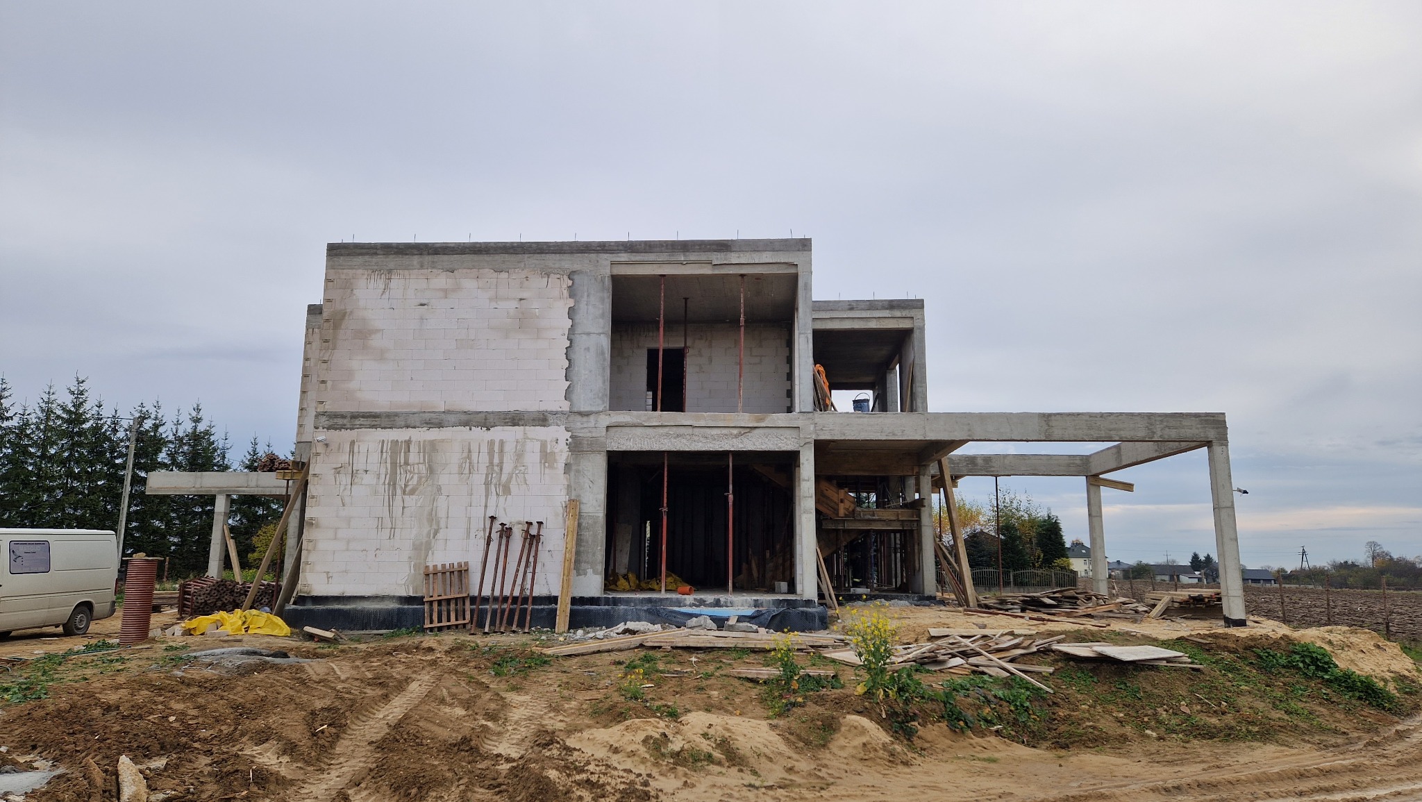Surowy stan dwukondygnacyjnego domu z betonu komórkowego w trakcie budowy, widoczne podpory i szalunki, teren budowy z ziemią i resztkami materiałów.