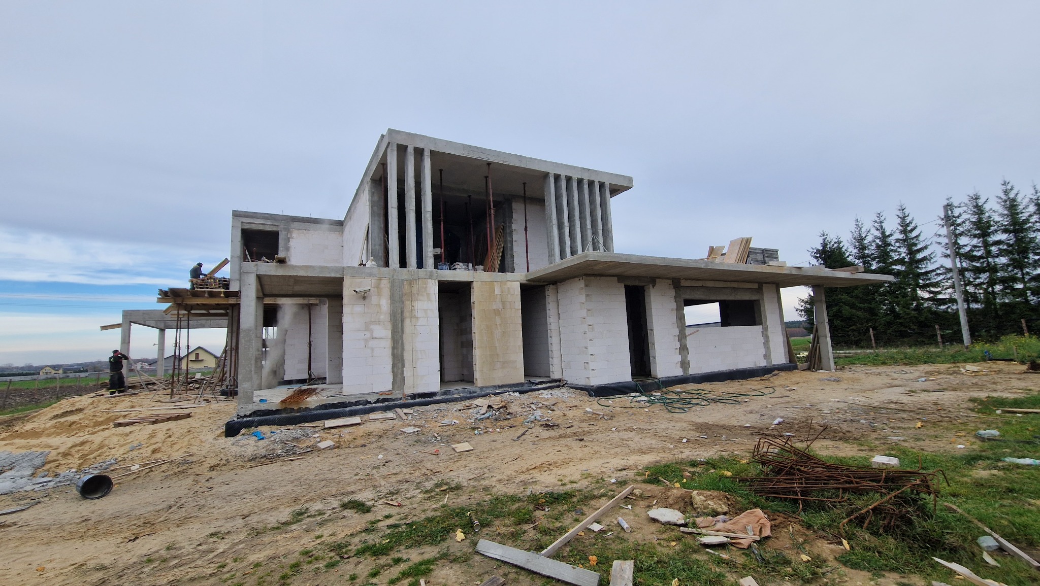 Surowy stan nowoczesnego domu z keramzytobetonu w trakcie budowy, widoczni pracownicy na rusztowaniu, teren wokół w stanie przygotowania, w tle las iglasty.