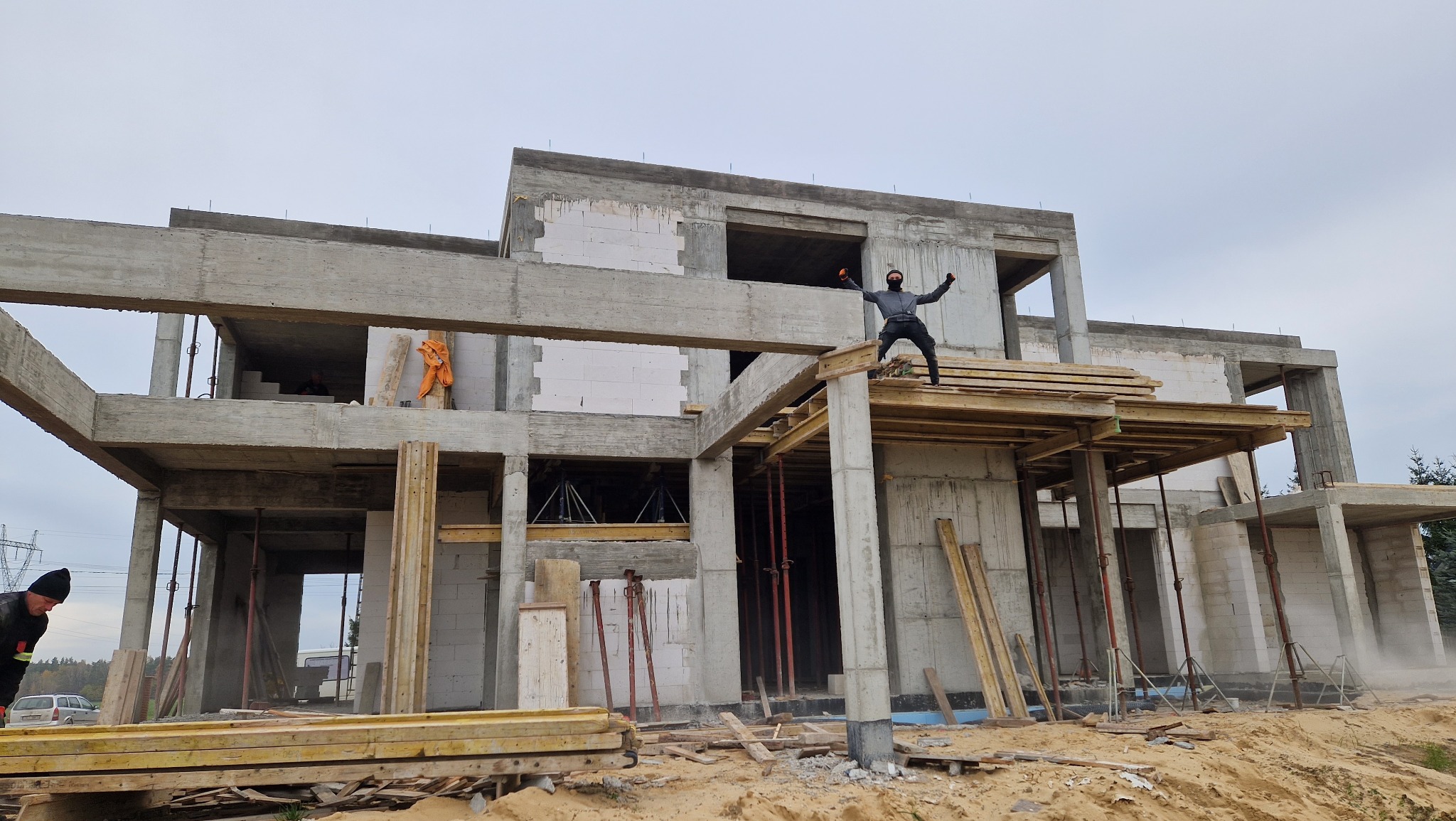 Surowy stan dwupiętrowego domu z betonu komórkowego w trakcie budowy, z osobą w masce stojącą na konstrukcji i rozkładającą ręce, widoczny zarys przyszłych okien i drzwi, drewniane podpory i szalunki.