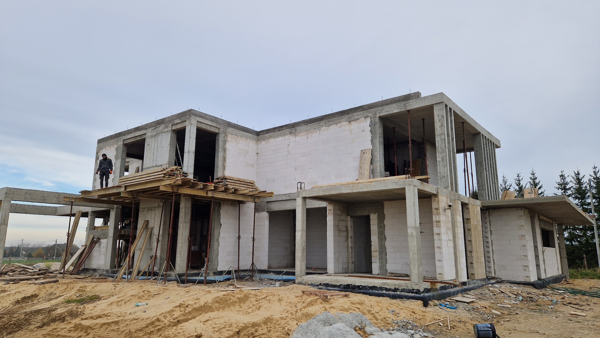 Niedokończony dom murowany z betonu komórkowego z widocznym zbrojeniem, deskowaniem i pracownikiem stojącym na rusztowaniu na tle pochmurnego nieba.