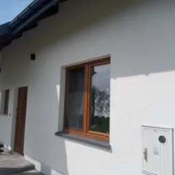 Boczny widok domu z białą elewacją o strukturze baranka, brązowe okno z firanką, widoczna skrzynka elektryczna i rynna, oraz fragment zadaszenia.