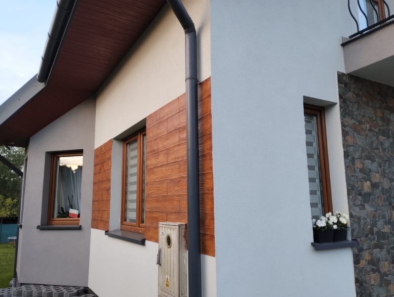 Fragment elewacji domu jednorodzinnego z elementami dekoracyjnymi imitującymi drewno i kamień, widoczne okna z roletami i doniczki z białymi kwiatami.