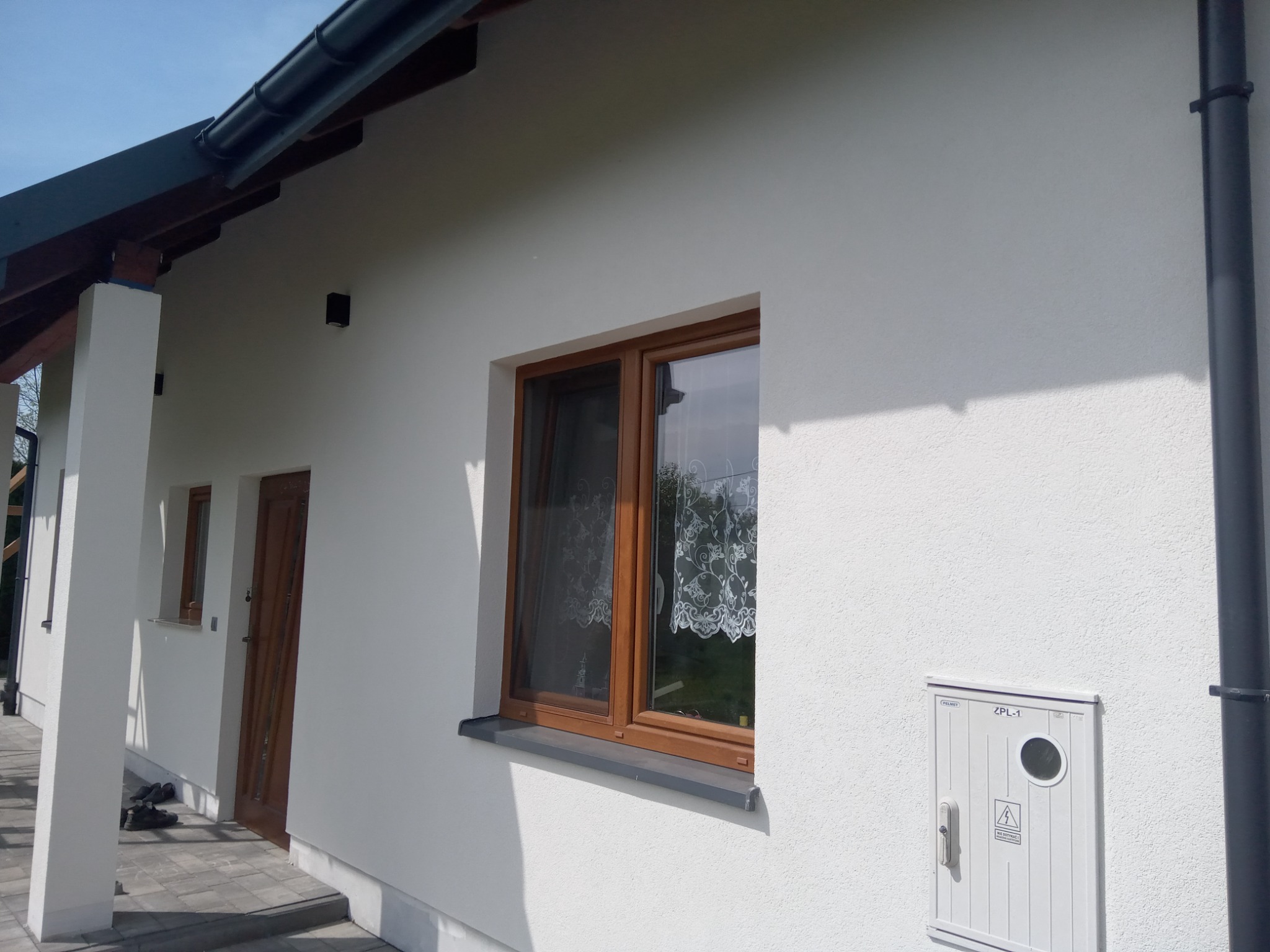 Boczny widok domu z białą elewacją o strukturze baranka, brązowe okno z firanką, widoczna skrzynka elektryczna i rynna, oraz fragment zadaszenia.