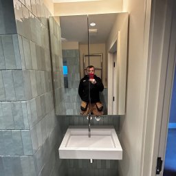 Adrian Walas - Nowoczesna łazienka z umywalką nablatową i lustrem w szafce, widoczne ułożone pionowo, błyszczące, szaro-niebieskie płytki oraz odbicie instalatora robiącego zdjęcie telefonem.