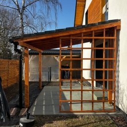 Adrian Walas - Drewniana konstrukcja zadaszenia tarasu w trakcie budowy, widoczne elementy kratownicy i słupy nośne, w tle dom jednorodzinny i osoba wewnątrz konstrukcji.