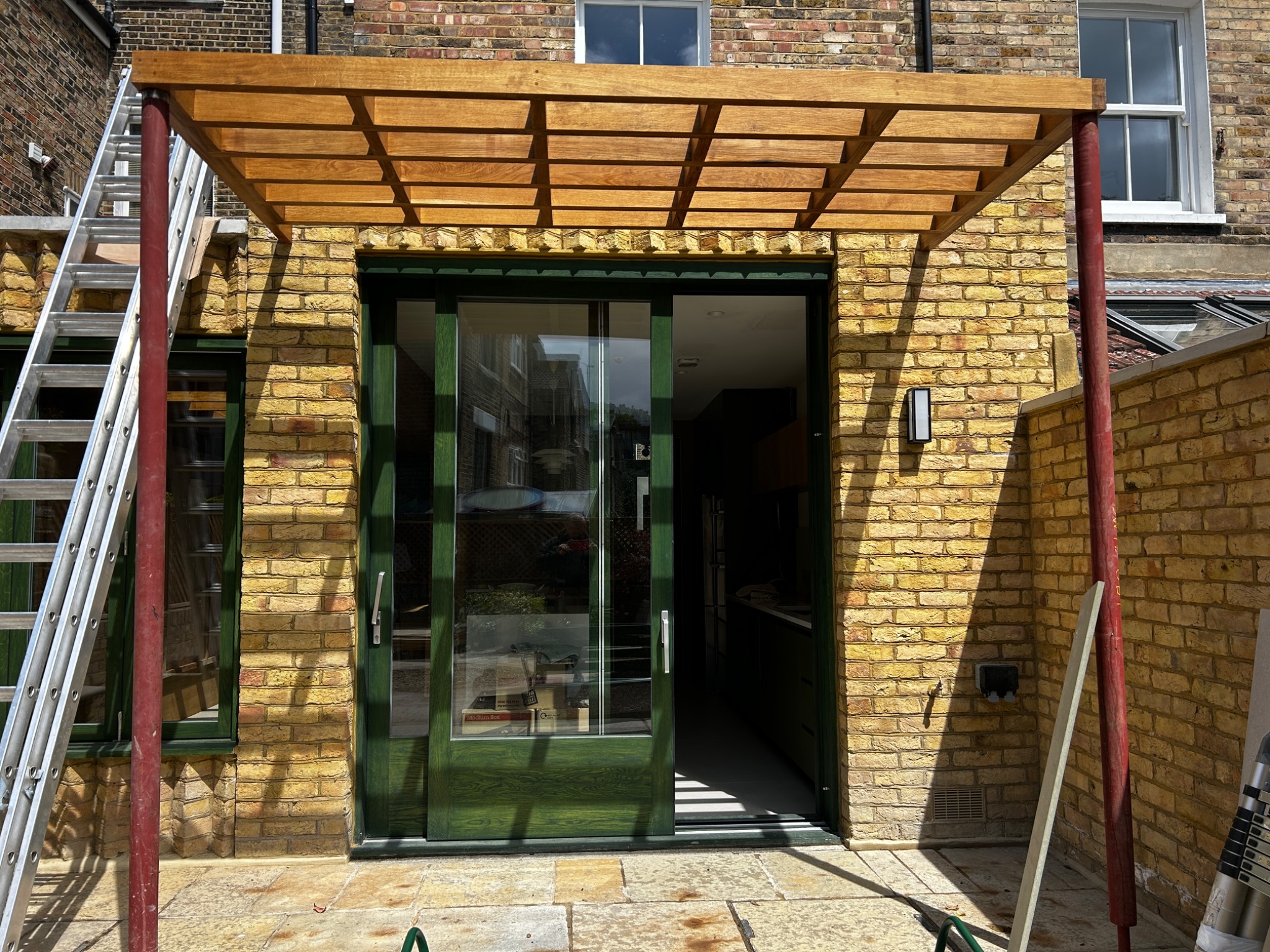 Drewniana pergola z kratownicą nad zielonymi, przesuwnymi drzwiami tarasowymi w domu z żółtej cegły, wsparta na metalowych, czerwonych słupach. Aluminiowa drabina oparta o ścianę. Widoczne...