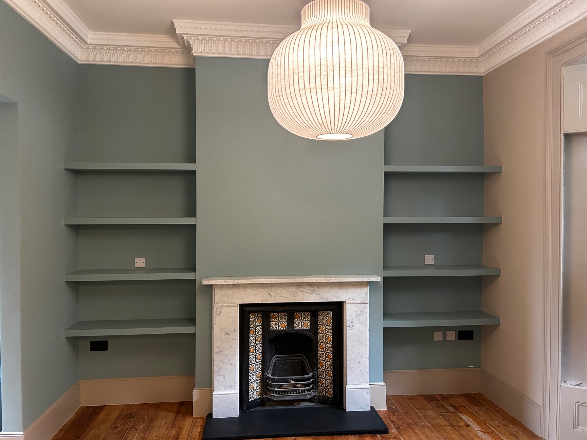 Wnętrze pokoju z zabudową wnęki: półki pomalowane na kolor ściany, marmurowy kominek z ozdobnymi kaflami i wisząca lampa z cienkich listewek.