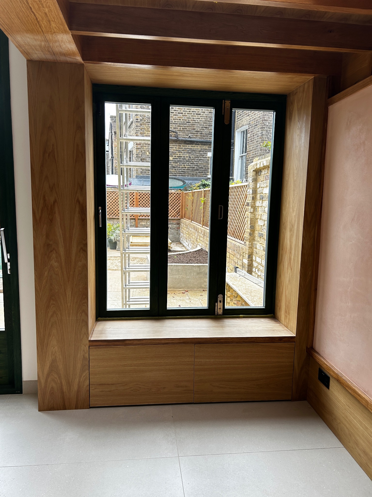 Drewniana zabudowa okienna z szafkami w dolnej części, widok na ogród z drabiny opartą o ogrodzenie panelowe w tle.