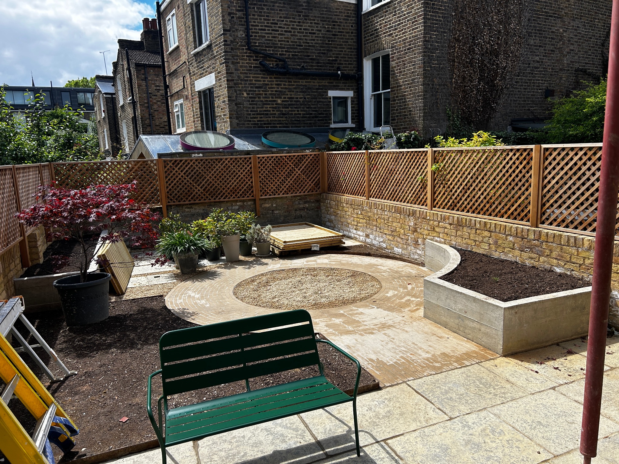 Aranżacja ogrodu w trakcie realizacji: drewniana kratownica ogrodzeniowa, wybrukowany okrąg z żwirowym wypełnieniem, podwyższona betonowa rabata, zielona metalowa ławka i drabina malarska.
