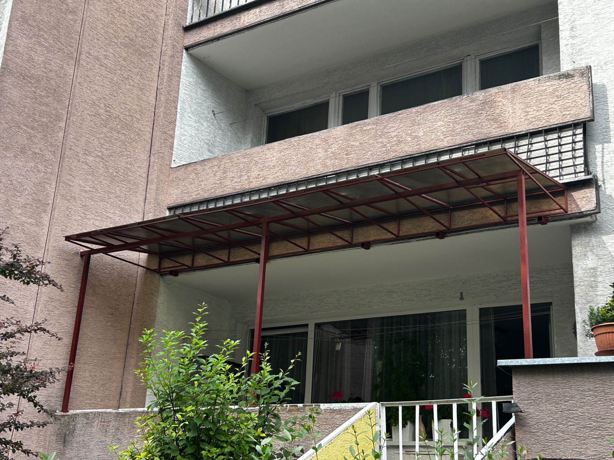 Metalowa konstrukcja z zadaszeniem, prawdopodobnie pergola, przymocowana do balkonu bloku mieszkalnego, widoczna z poziomu ogrodu z zielenią.
