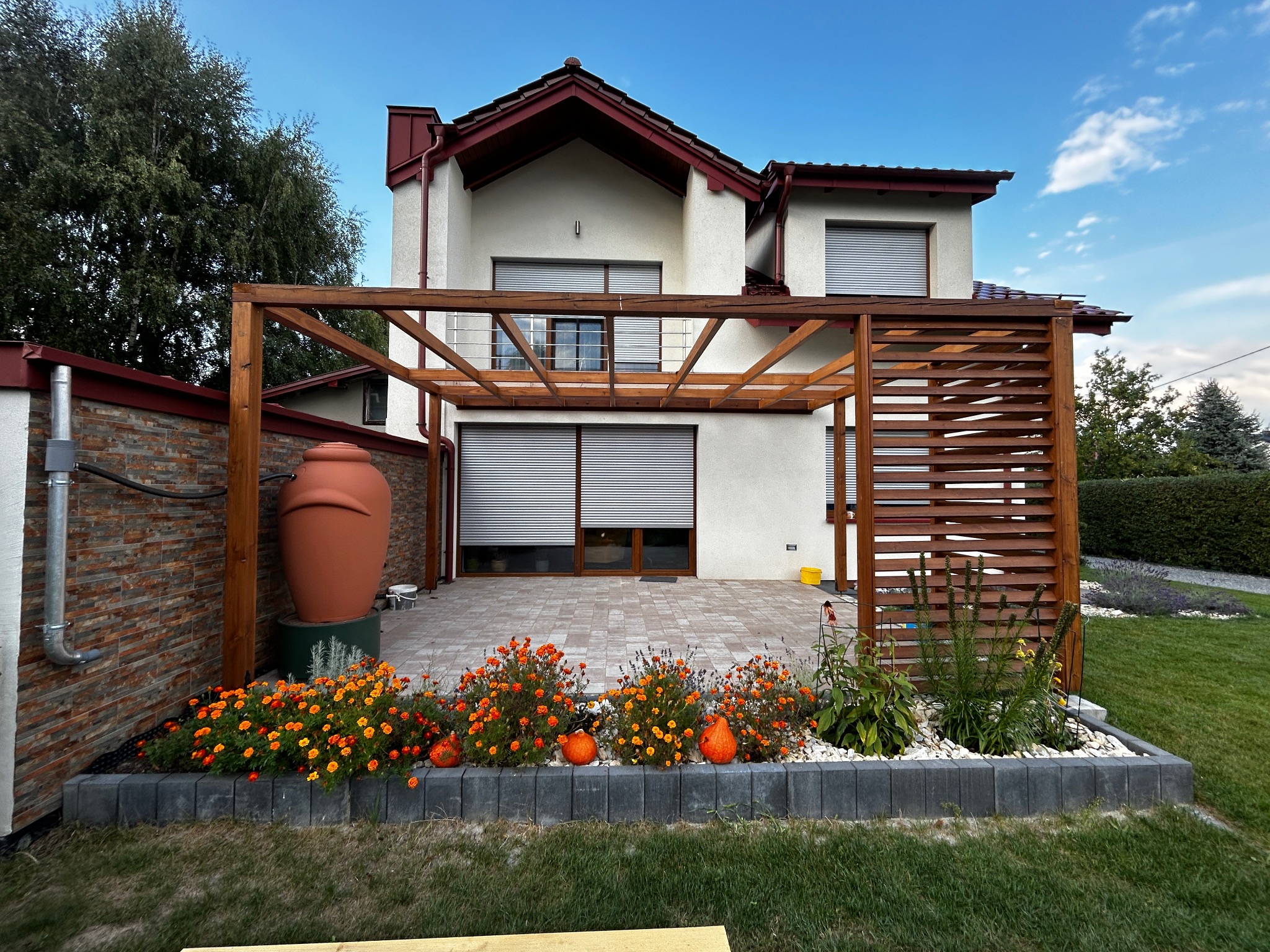 Drewniana pergola z poziomymi listwami, przylegająca do domu z zaciągniętymi roletami, obok duży zbiornik na deszczówkę i rabata z pomarańczowymi aksamitkami oraz dekoracyjnymi dyniami.