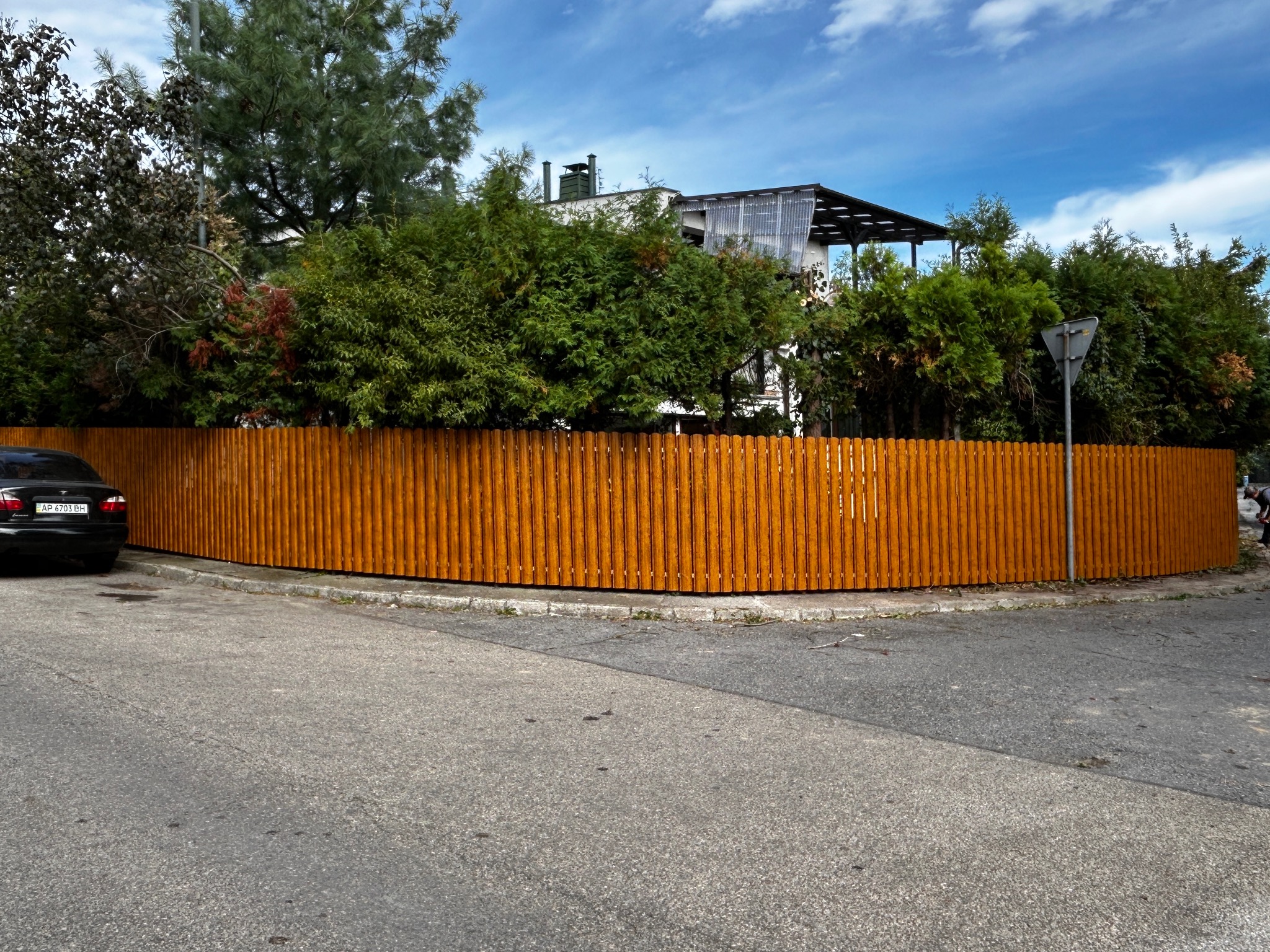 Nowoczesne ogrodzenie z pionowych desek w kolorze ciepłego brązu, otaczające posesję z bujną zielenią, widoczne z poziomu ulicy z zaparkowanym czarnym samochodem.