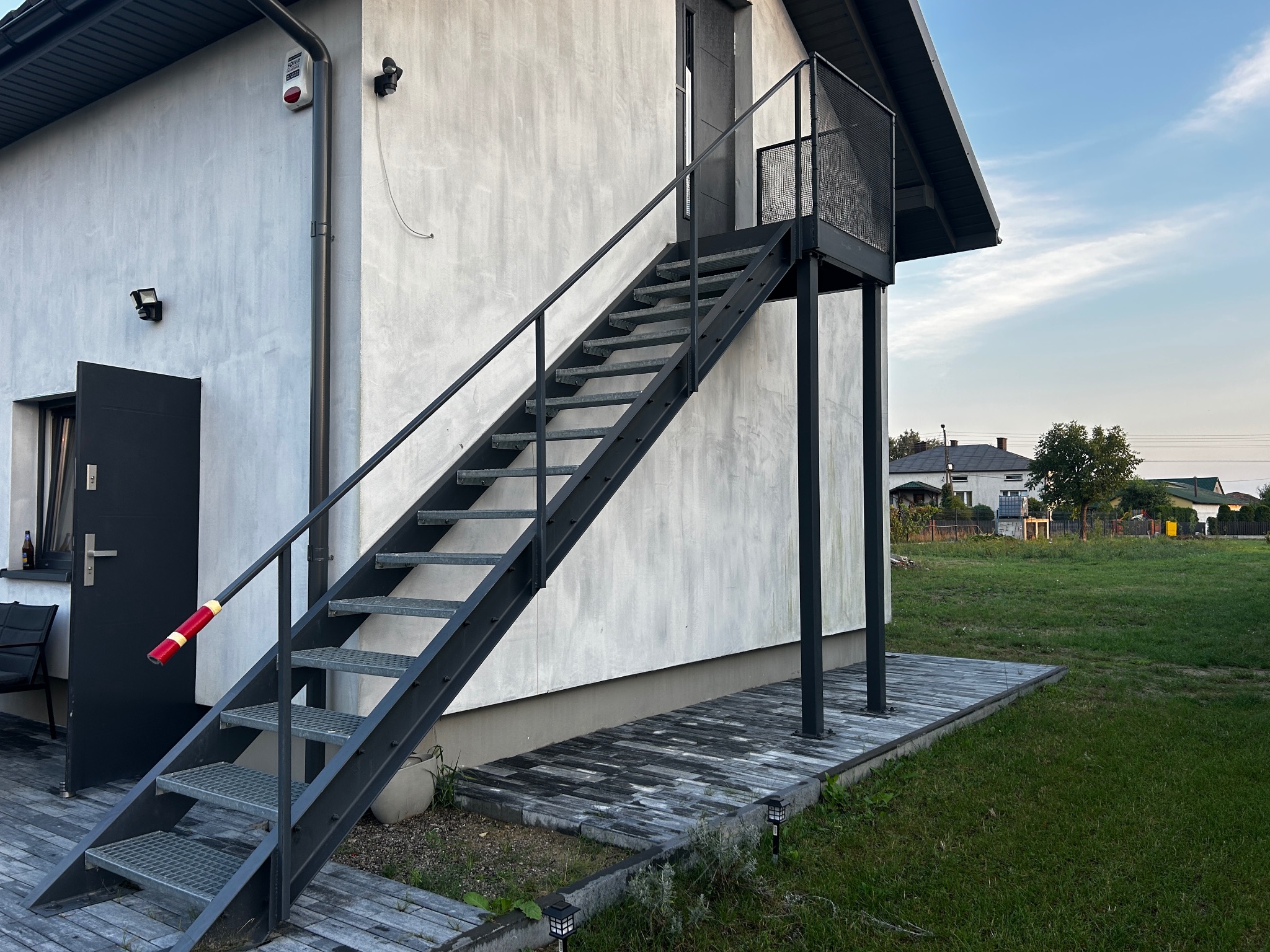 Zewnętrzne schody metalowe z platformą prowadzące do drzwi na piętrze, z poręczą z czerwoną taśmą, zamontowane przy białym budynku z drewnianym tarasem.