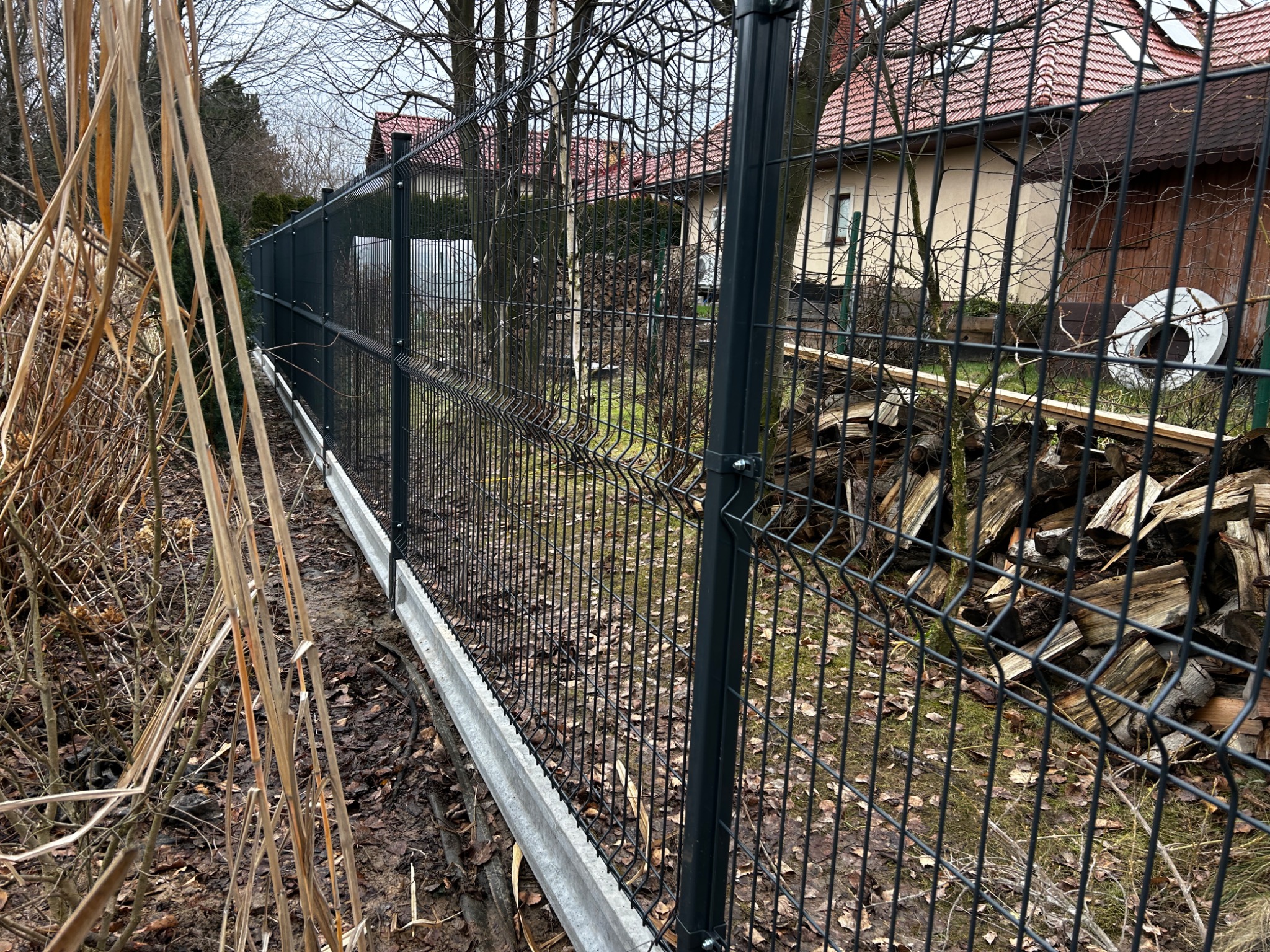Nowoczesne ogrodzenie panelowe w kolorze grafitowym, z betonową podmurówką, oddzielające posesję od zarośli i sąsiedniego domu z zapasem drewna.