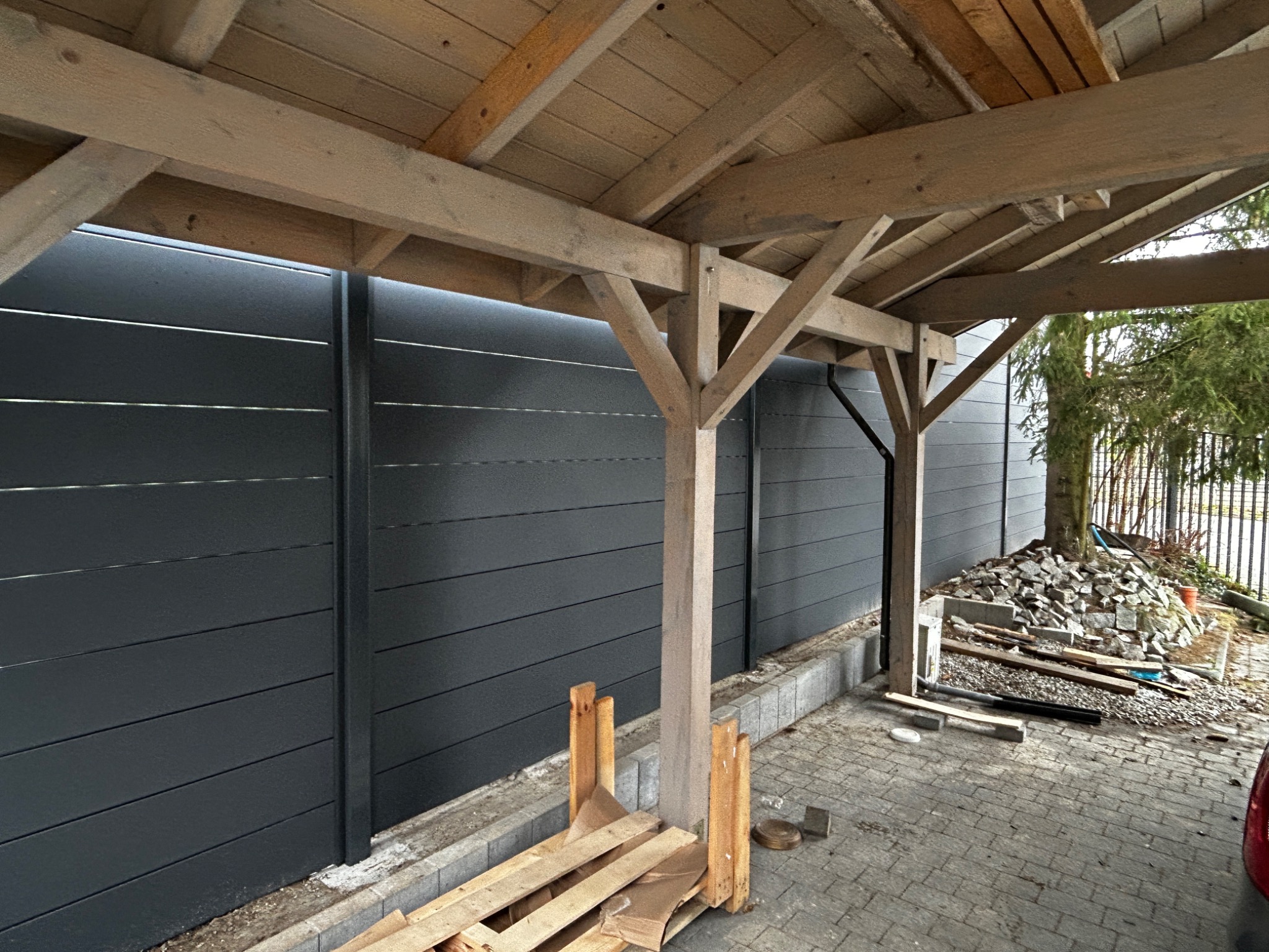 Widok spod drewnianej pergoli w trakcie budowy, z widocznym fragmentem ciemnoszarego, poziomego ogrodzenia panelowego, paletą i stertą kamieni w tle.