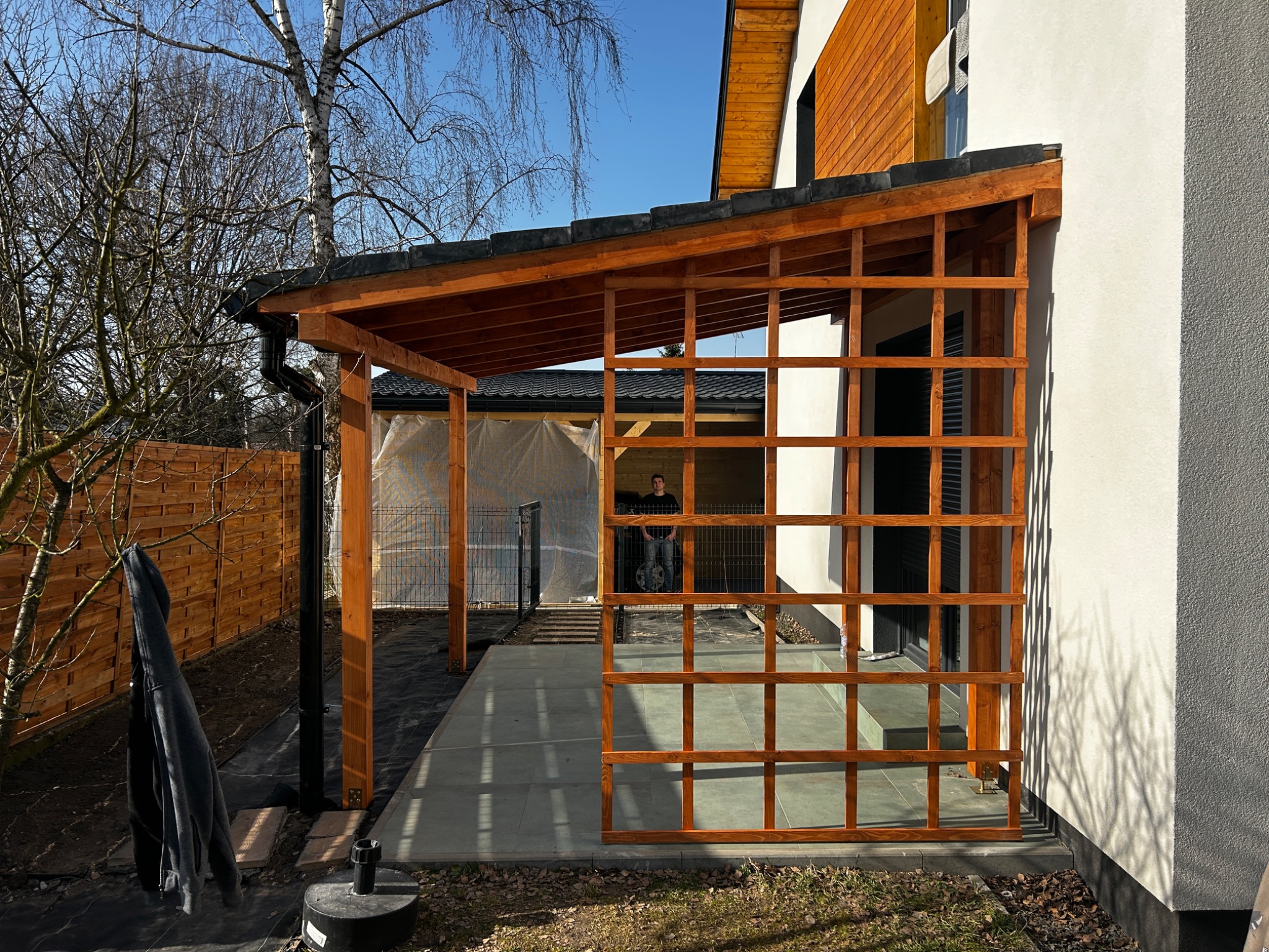 Drewniana konstrukcja zadaszenia tarasu w trakcie budowy, widoczne elementy kratownicy i słupy nośne, w tle dom jednorodzinny i osoba wewnątrz konstrukcji.