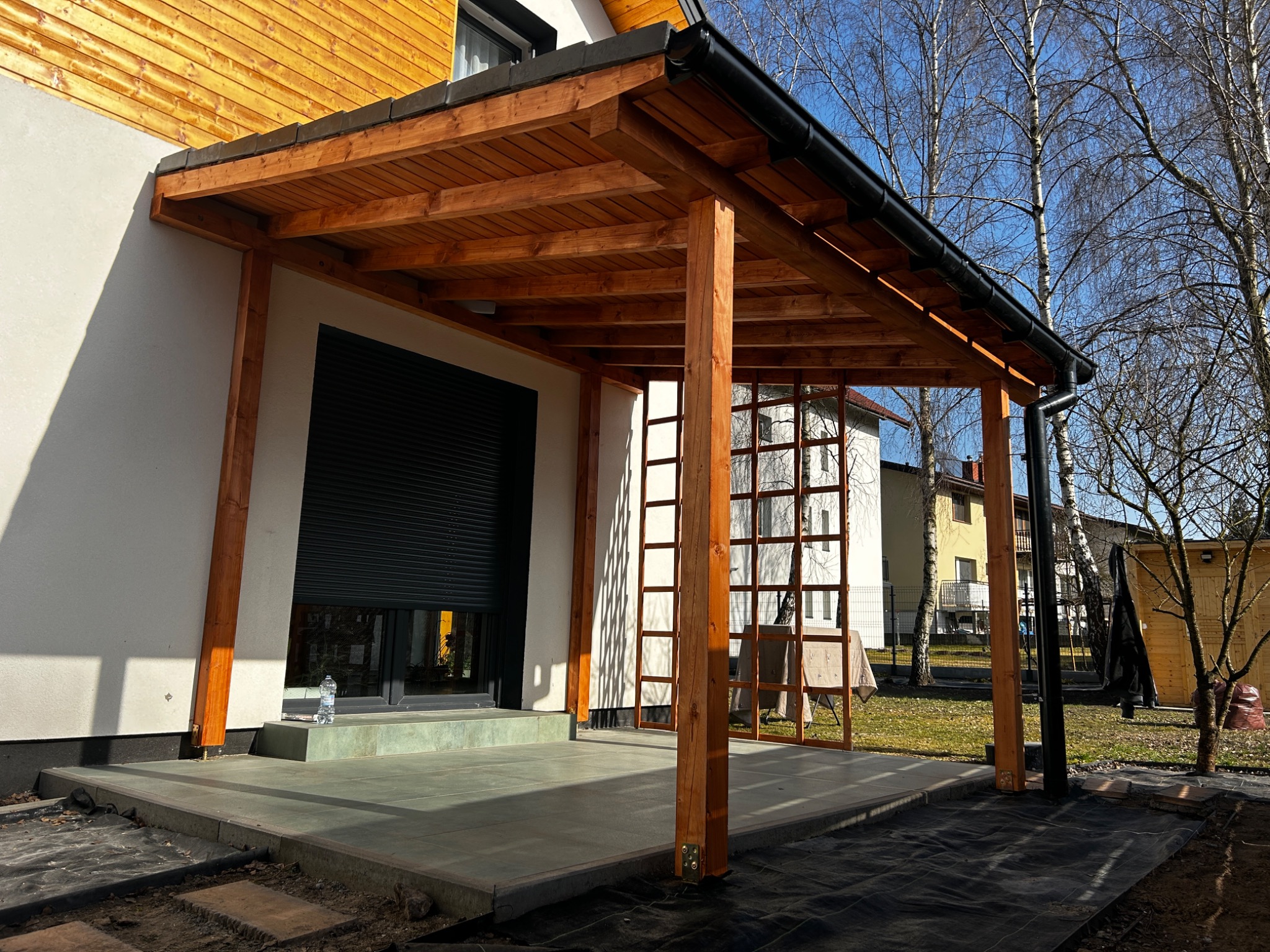 Drewniana pergola z czarną rynną przylegająca do jasnego budynku z roletą, widok na betonowy taras i przygotowany teren pod ogród.