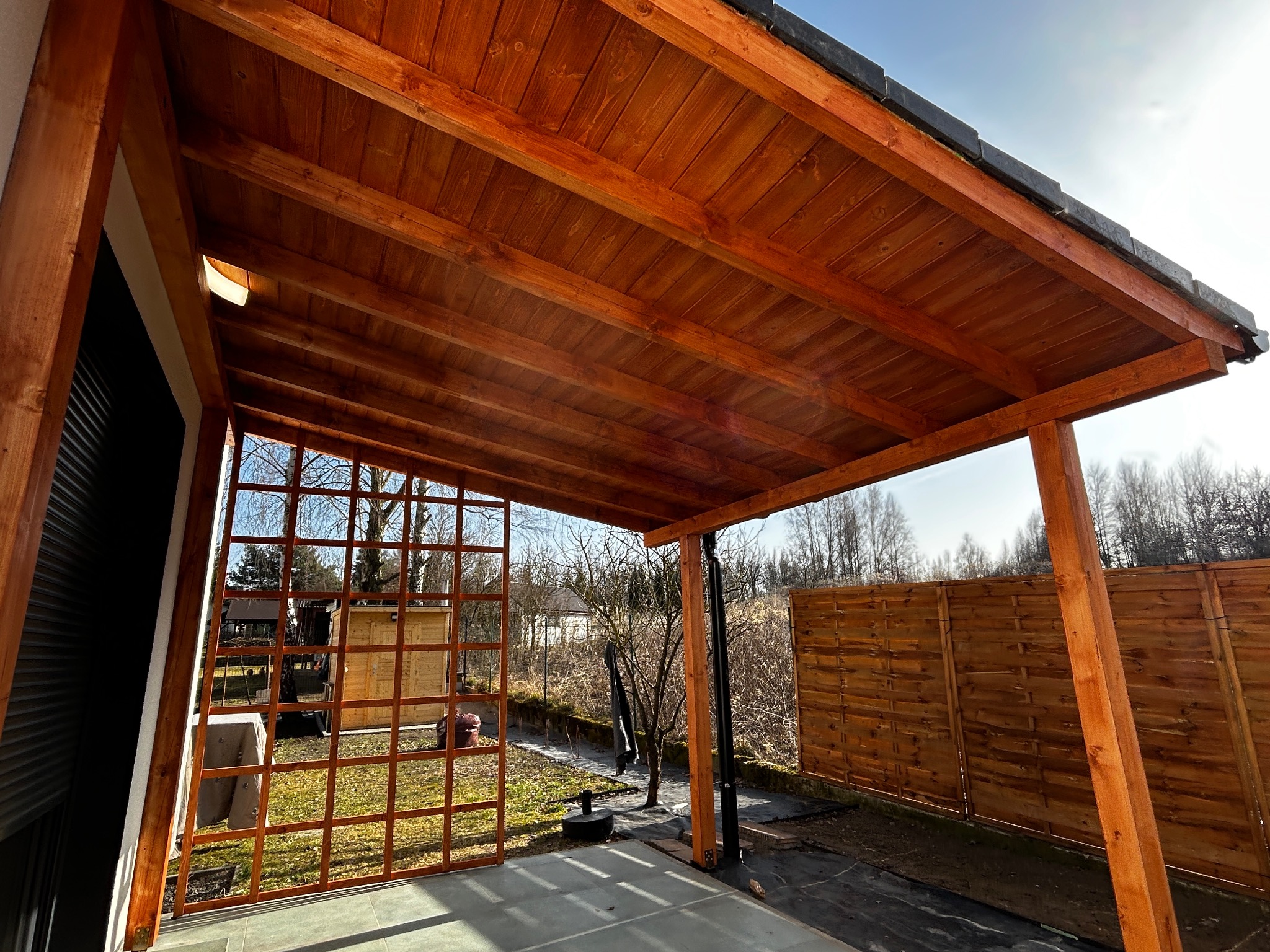 Drewniana pergola z zadaszeniem z desek, widok od wewnątrz, z kratownicą po lewej stronie i fragmentem drewnianego ogrodzenia w tle.
