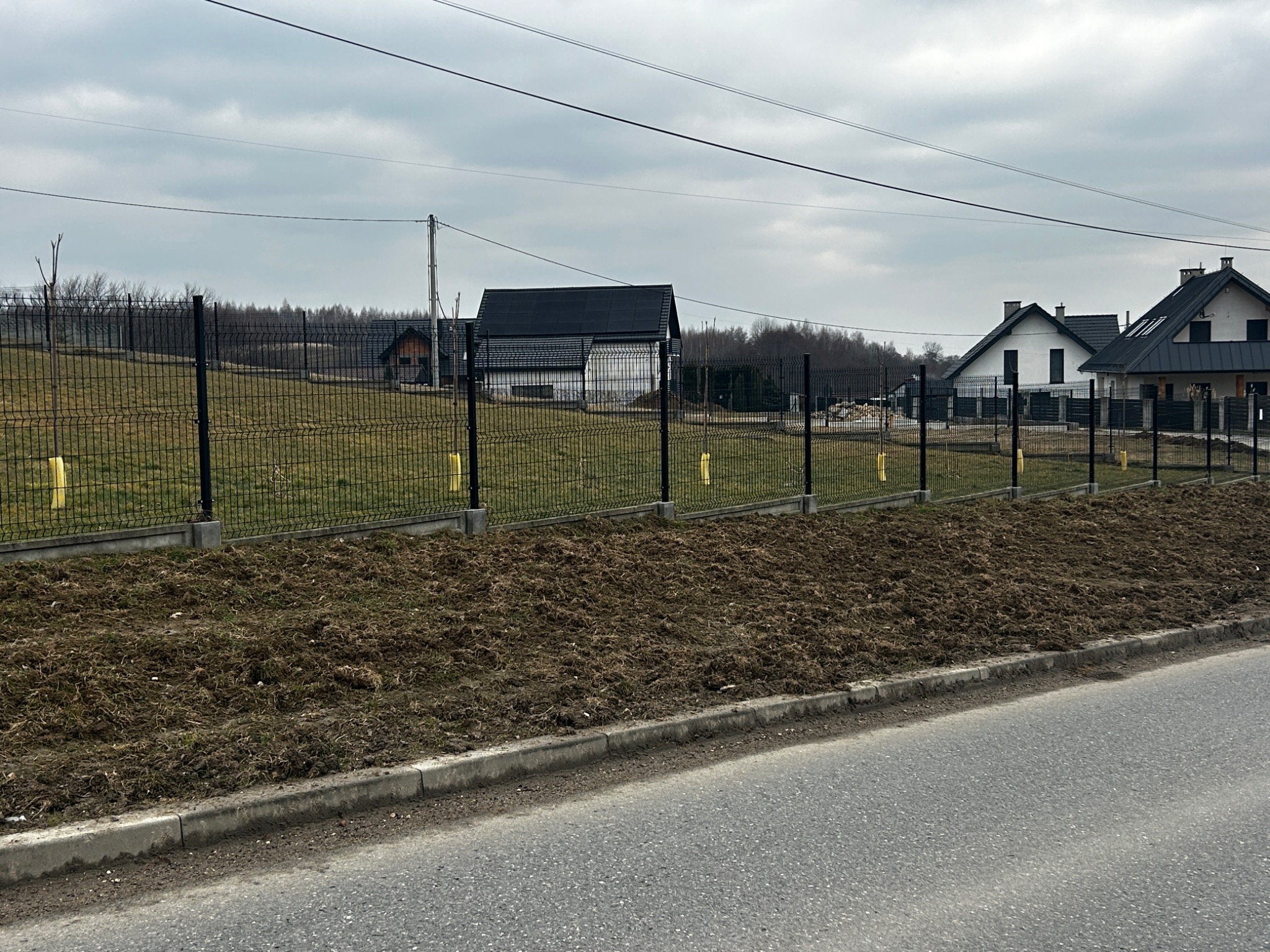 Nowo postawione czarne ogrodzenie panelowe z żółtymi słupkami tymczasowymi na tle domów jednorodzinnych i pochmurnego nieba.