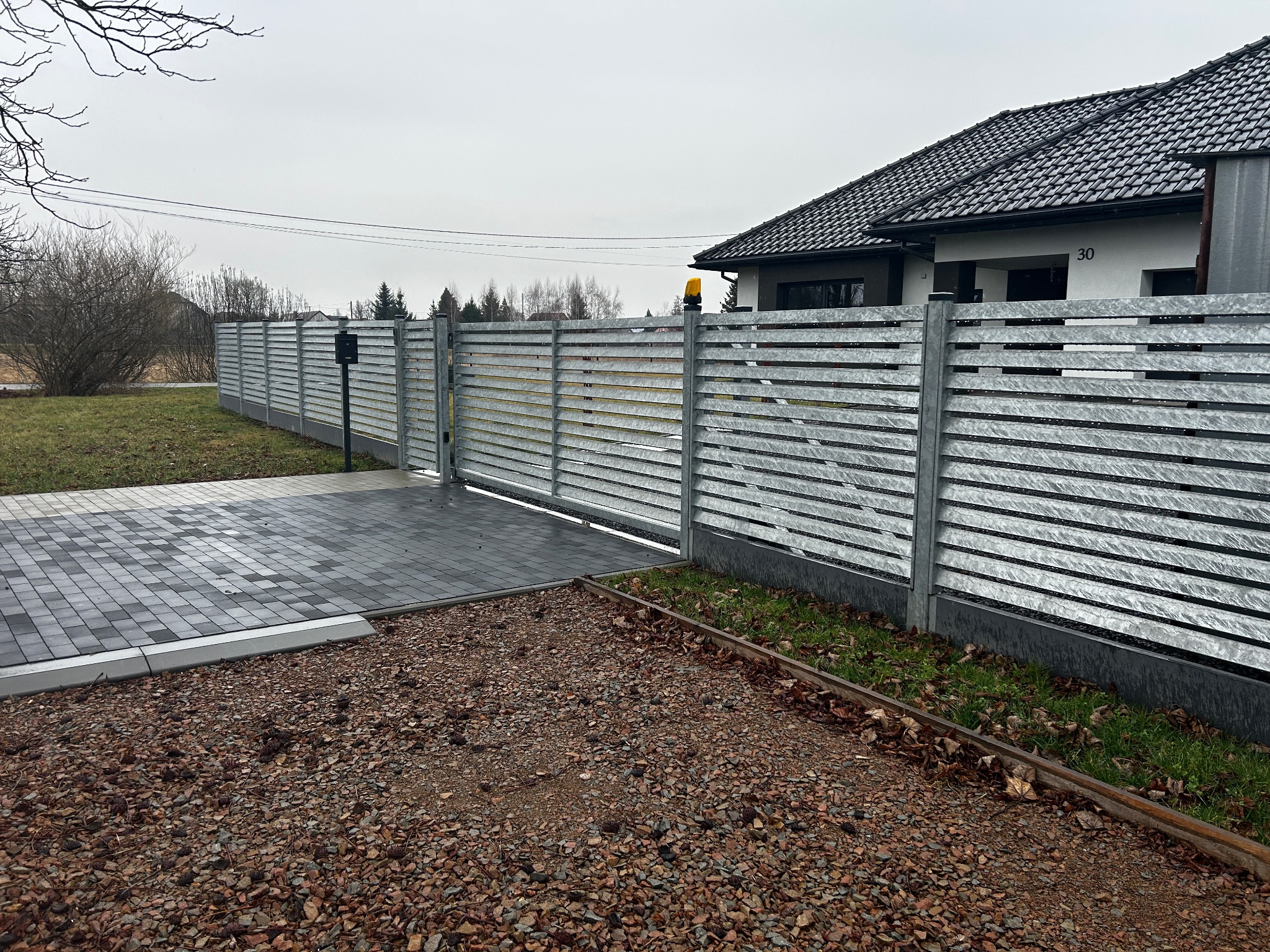 Nowoczesne ogrodzenie posesji z poziomych, metalowych profili, brama wjazdowa z automatyką, kostka brukowa na podjeździe, dom jednorodzinny w tle.