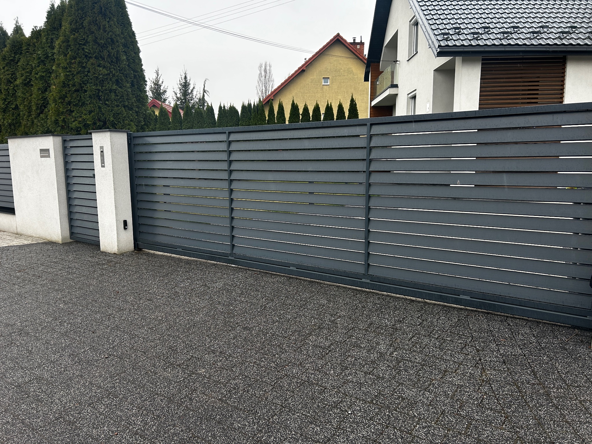 Nowoczesna, przesuwna brama posesyjna z poziomych, szarych profili, zamontowana między dwoma białymi, otynkowanymi słupkami z wideodomofonem i skrzynką na listy, na tle kostki brukowej i zabudowy...