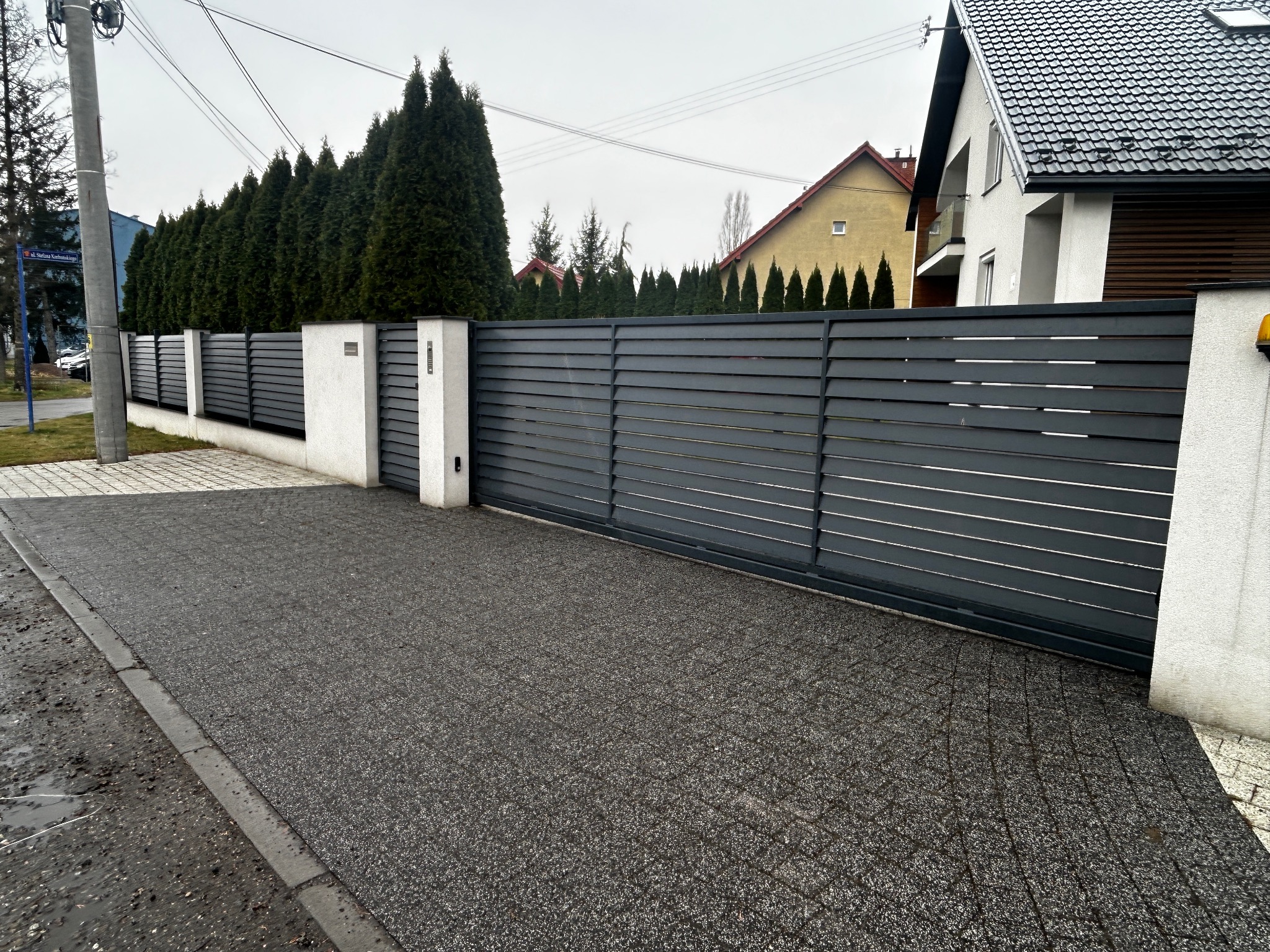 Nowoczesne, poziome ogrodzenie z szarych paneli z białymi słupkami, na tle rzędu wysokich, zielonych tuj i domów jednorodzinnych.
