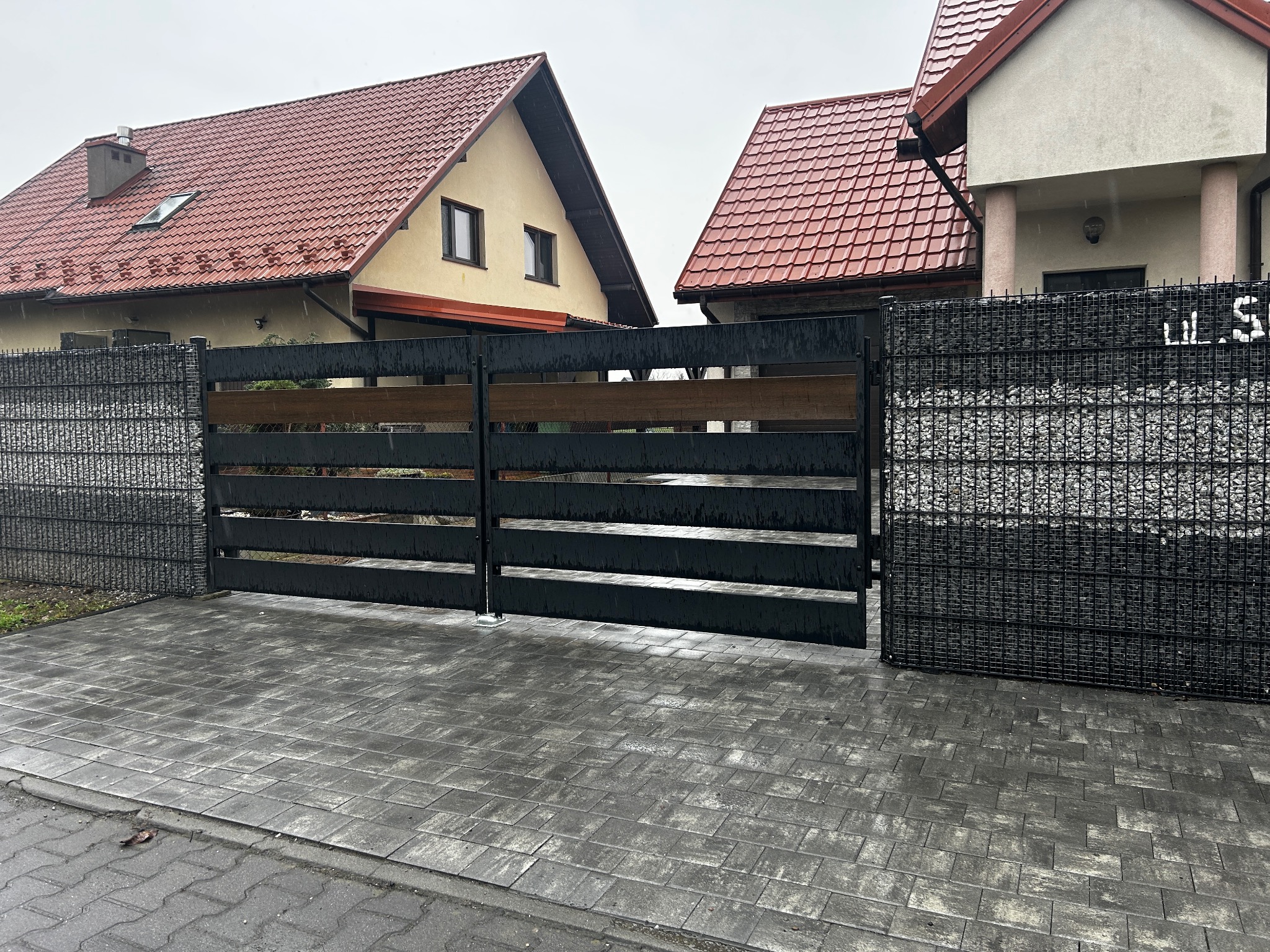 Nowoczesna brama wjazdowa z czarnych, poziomych paneli z drewnianymi akcentami, osadzona w gabionowym ogrodzeniu z szarego kamienia, na tle dwóch domów z czerwonymi dachami w deszczowy dzień.