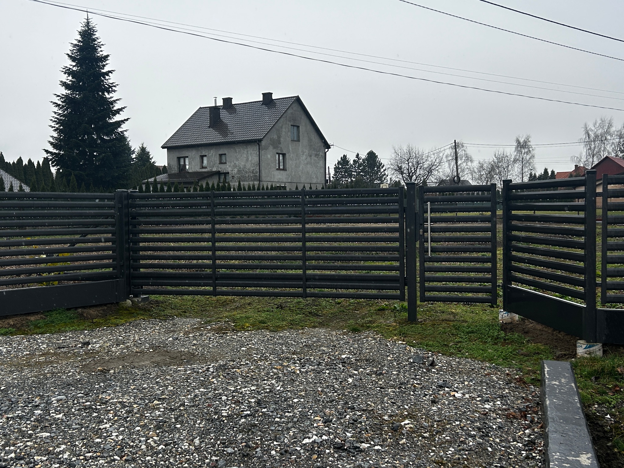 Nowoczesne, poziome ogrodzenie z czarnych paneli metalowych z otwartą bramą wjazdową, widok na dom i drzewa w pochmurny dzień.