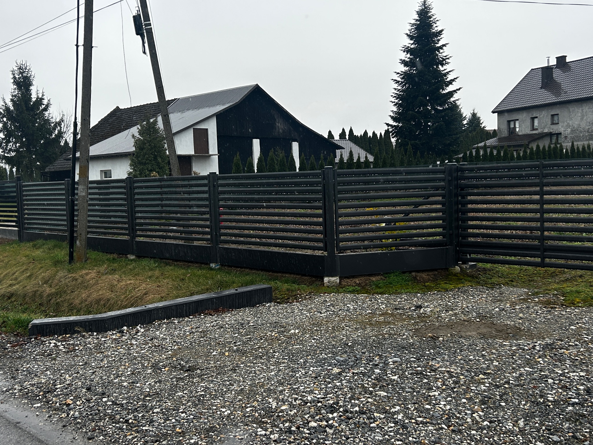 Nowoczesne ogrodzenie panelowe z poziomych, czarnych listew, oddzielające posesję z domami i iglakami od żwirowego podłoża w pochmurny dzień.
