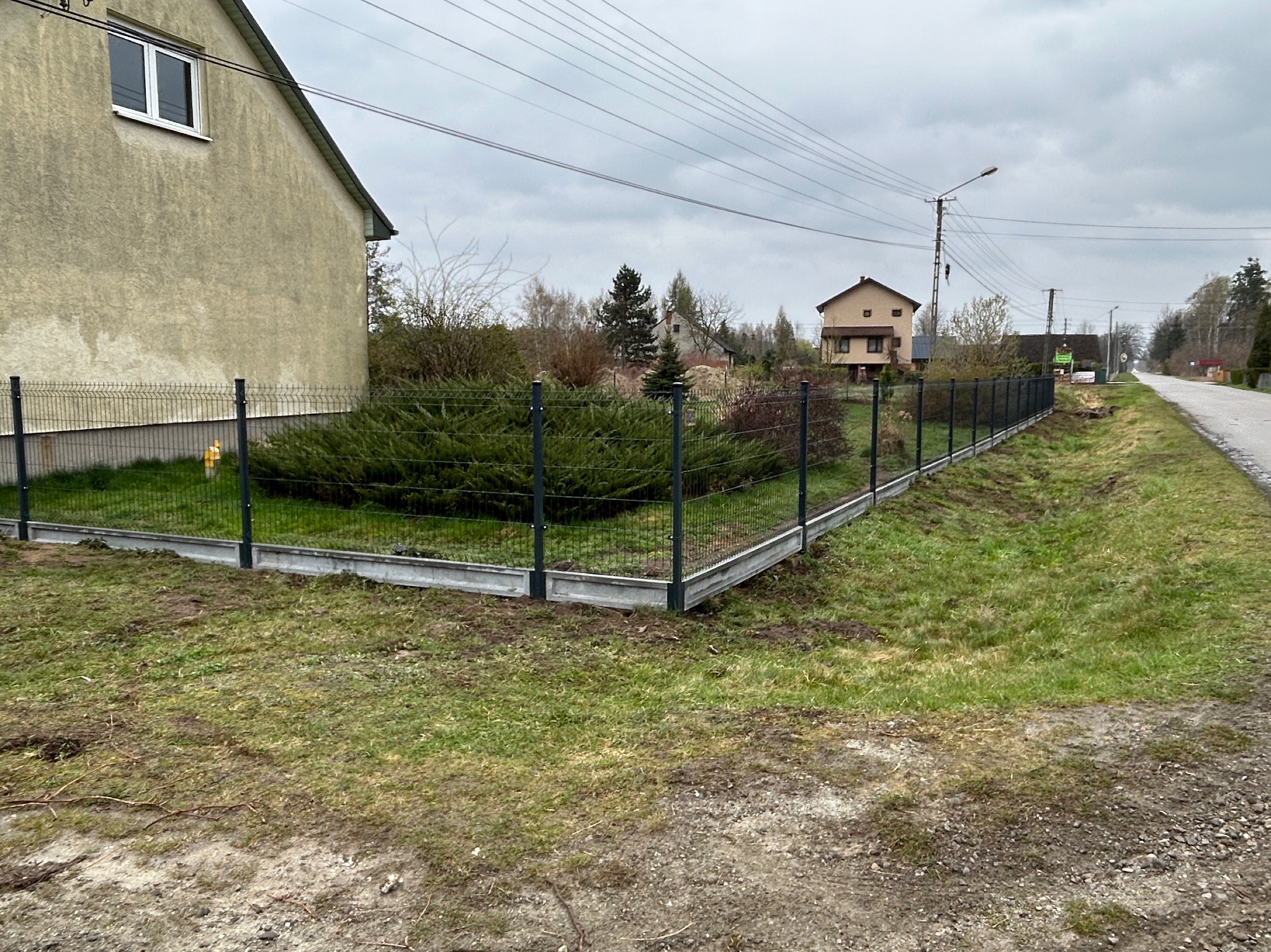 Fragment nowo postawionego, czarnego ogrodzenia panelowego z betonową podmurówką, widoczne krzewy i trawa, w tle dom jednorodzinny i słupy elektryczne, pochmurne niebo.