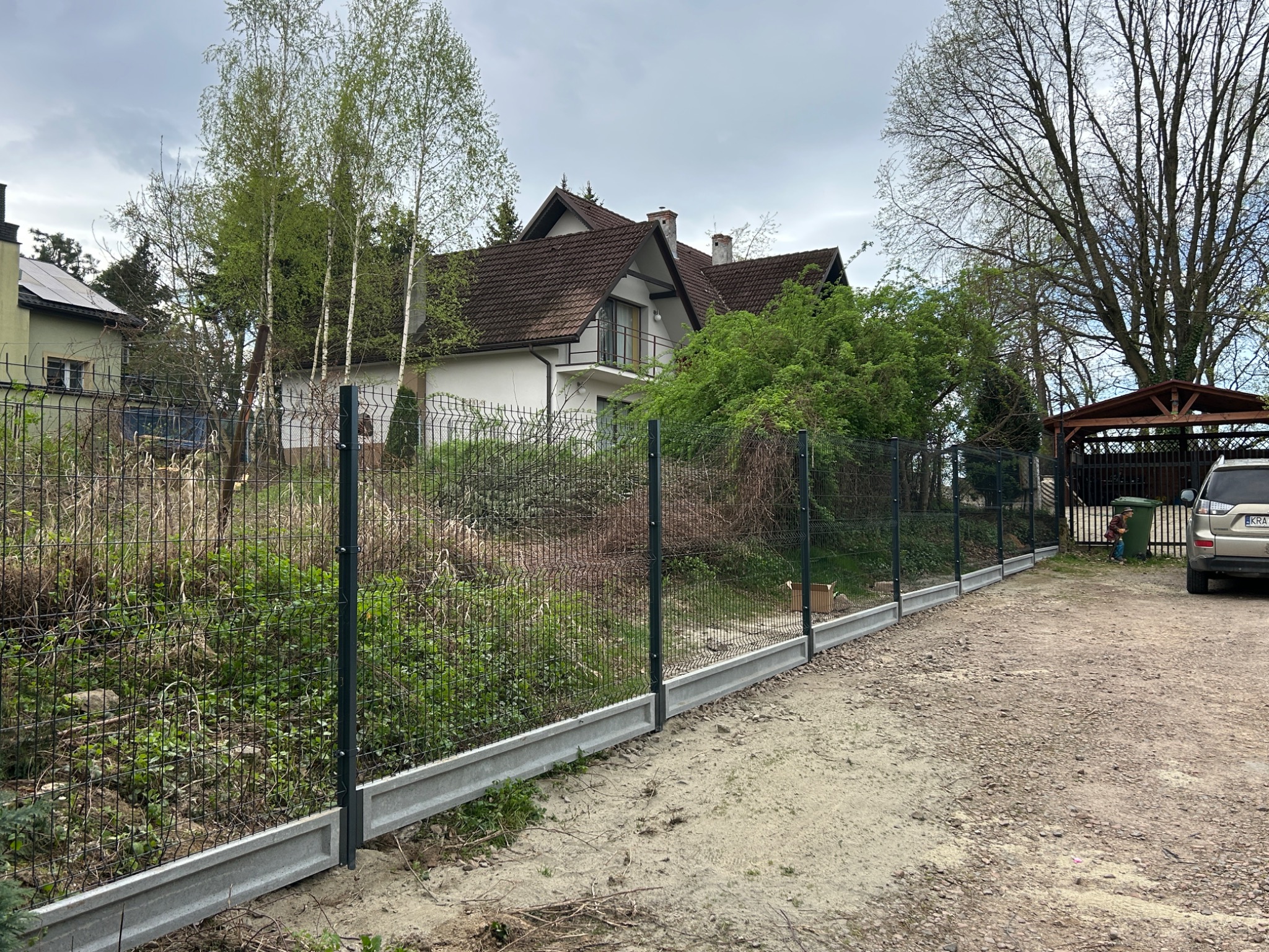 Nowe ogrodzenie panelowe z podmurówką betonową na posesji, widok z zewnątrz. Szara siatka i słupki na tle zieleni i domu z brązowym dachem w tle. Drobny żwir na podłożu.