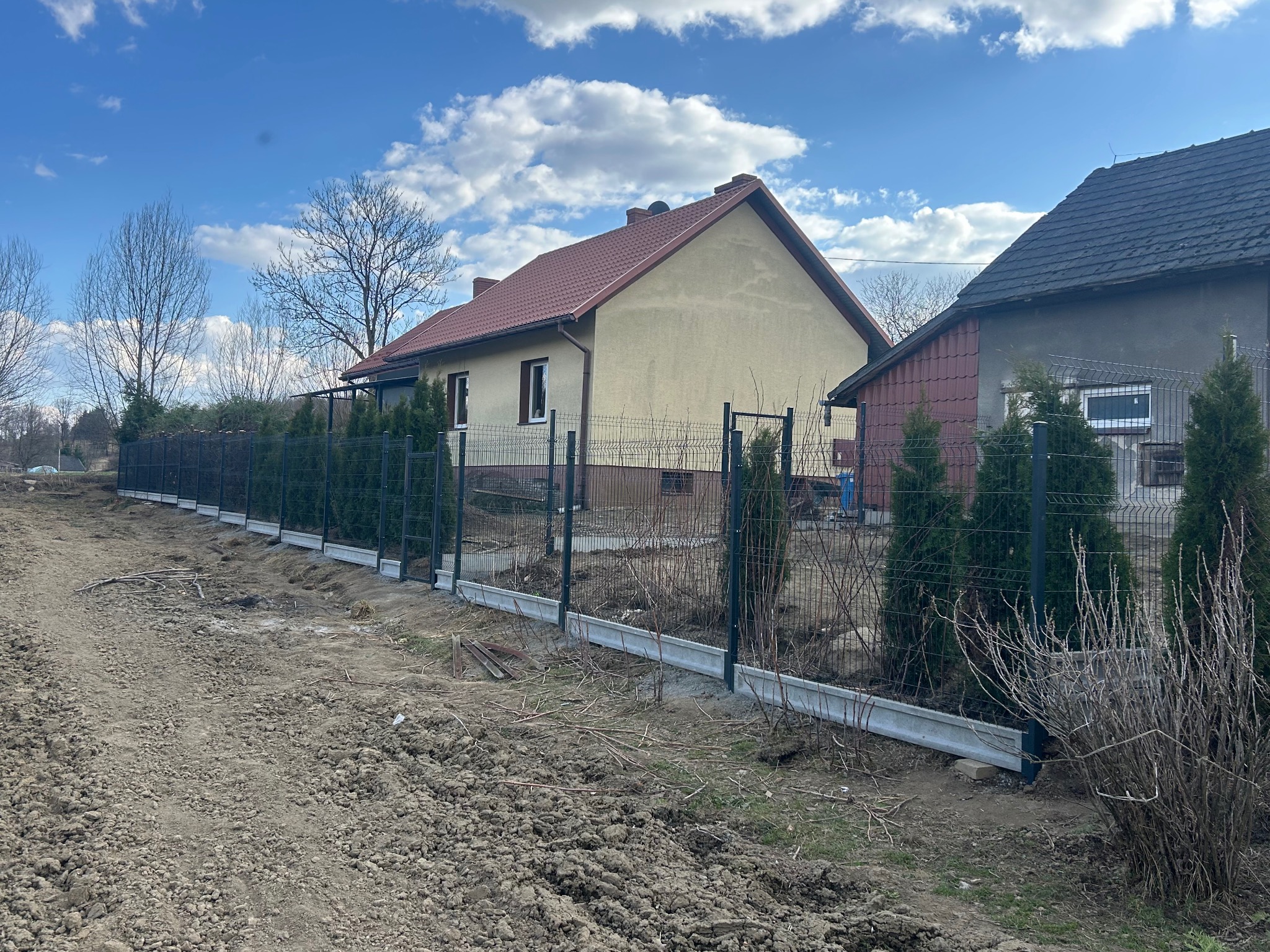 Nowoczesne ogrodzenie panelowe w kolorze grafitowym, z podmurówką betonową, oddzielające posesję z domem od sąsiedniej działki. Widok w słoneczny dzień.