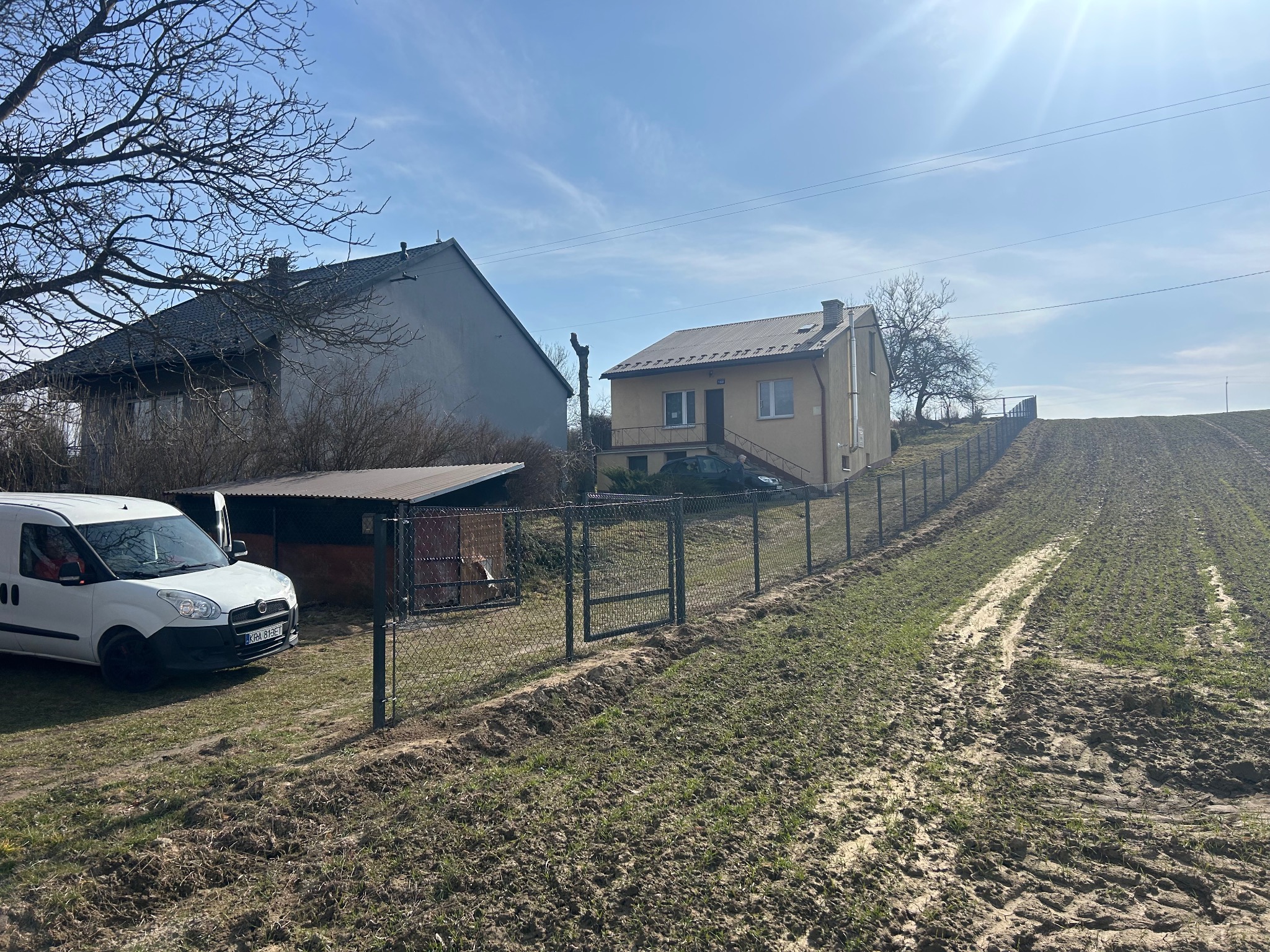 Montaż ogrodzenia z siatki na nierównym terenie, widoczny dom i samochód dostawczy. Słoneczny dzień, świeżo posiana trawa. Brama w ogrodzeniu.